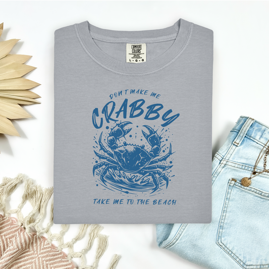 Comfort Colors 1717 "Don't Make Me Crabby Take Me To The Beach" t shirt with a crab splashing in water in a one color graphic. Picture on a flay lay mockup with pair of blue jeans and blanket and a wicker sun shaped accessory.