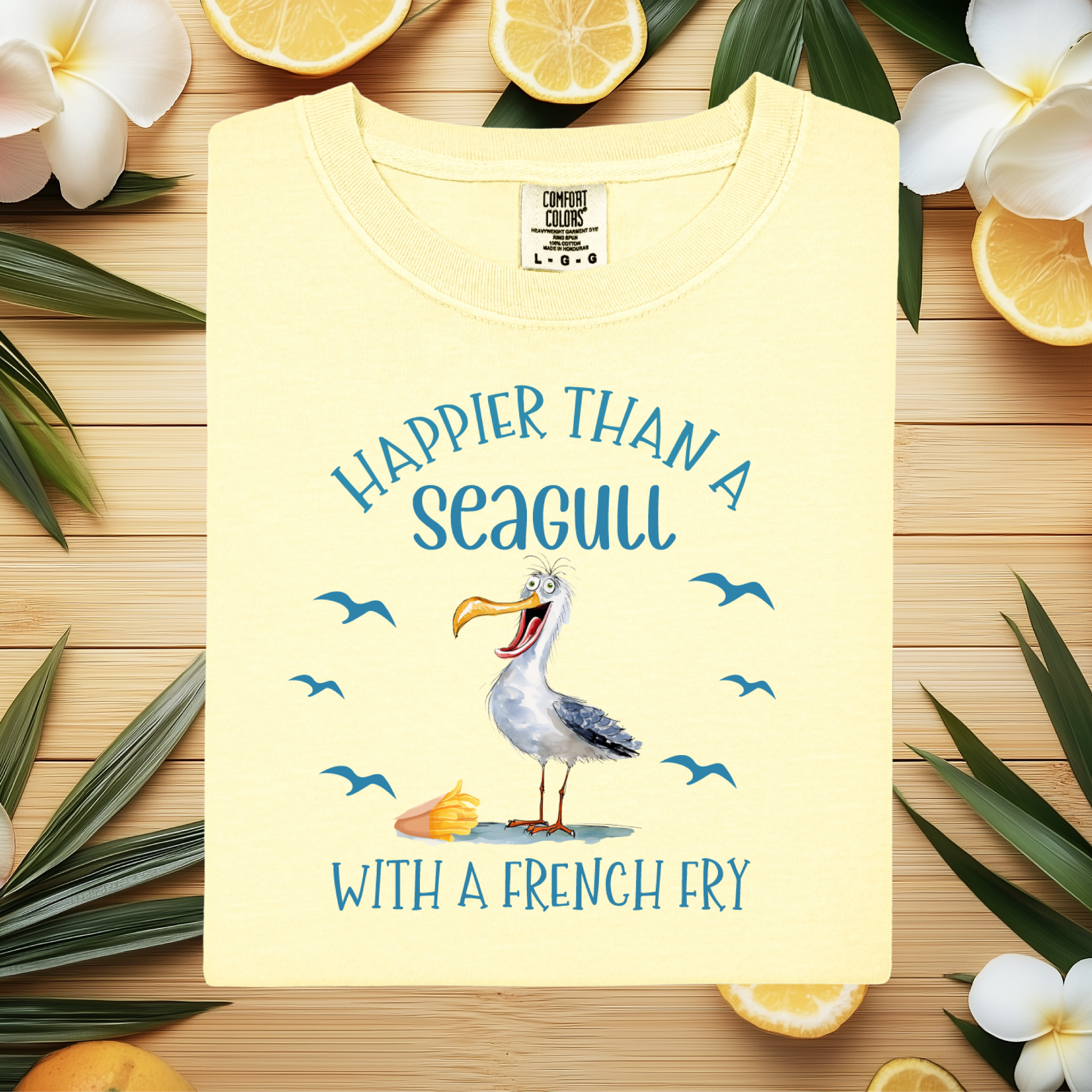 Yellow t-shirt with a seagull graphic and text on a wooden surface with lemons and flowers.