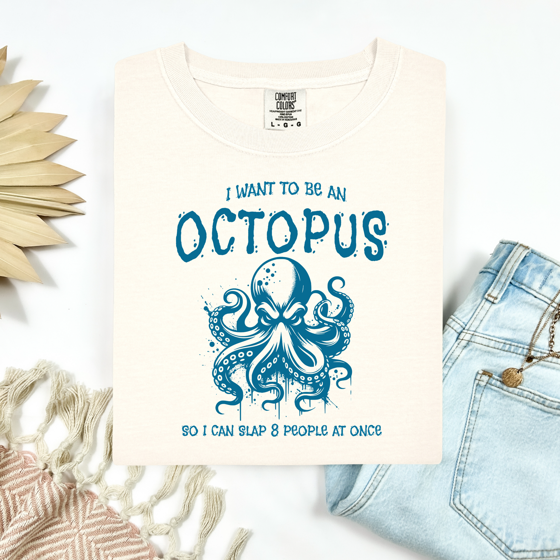 Comfort Colors 1717 "I Want To Be On Octopus So I Can Slap 8 People At Once" t shirt with an angry octopus one color graphic. Picture on a flay lay mockup with pair of blue jeans and blanket and a wicker sun shaped accessory.