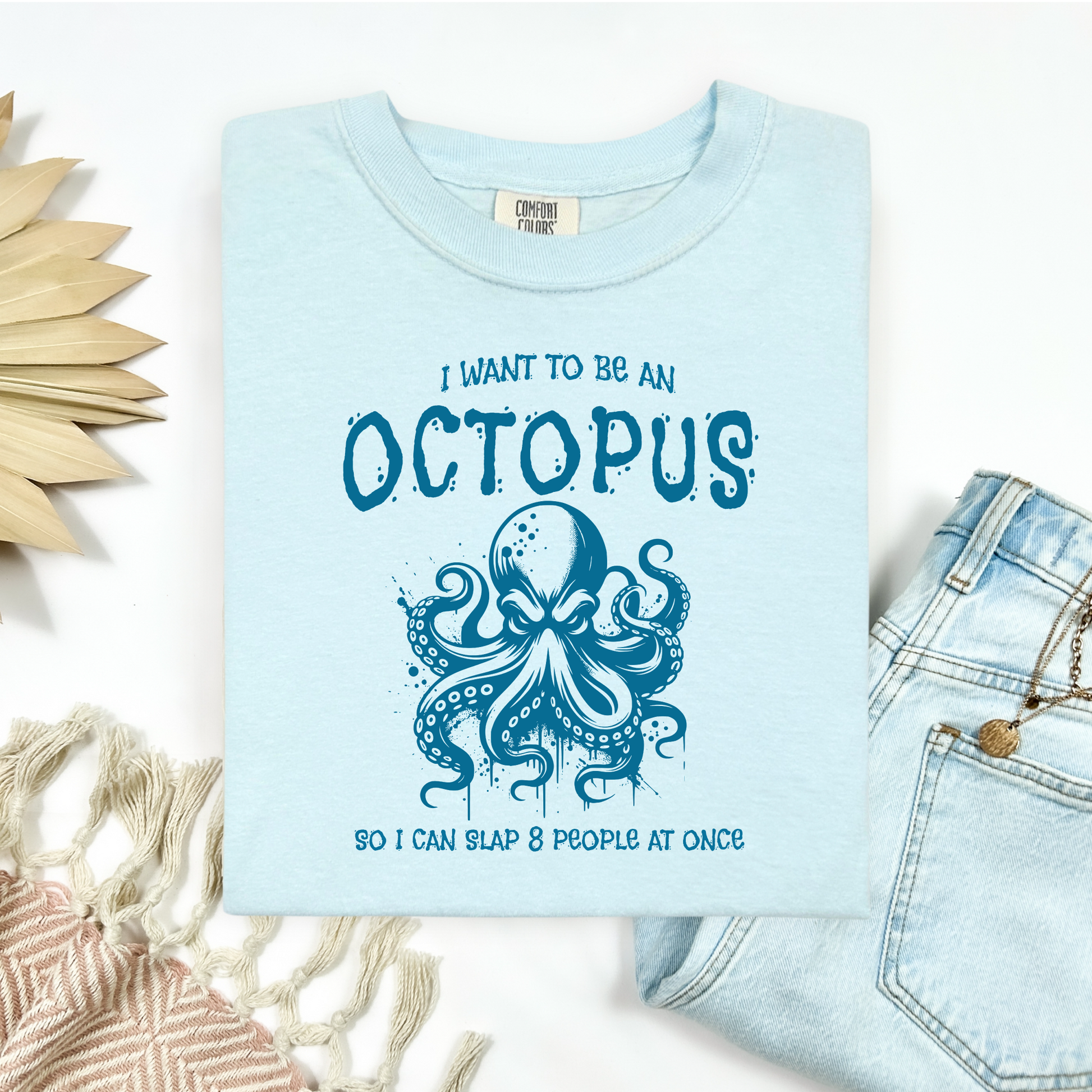 Comfort Colors 1717 "I Want To Be On Octopus So I Can Slap 8 People At Once" t shirt with an angry octopus one color graphic. Picture on a flay lay mockup with pair of blue jeans and blanket and a wicker sun shaped accessory.