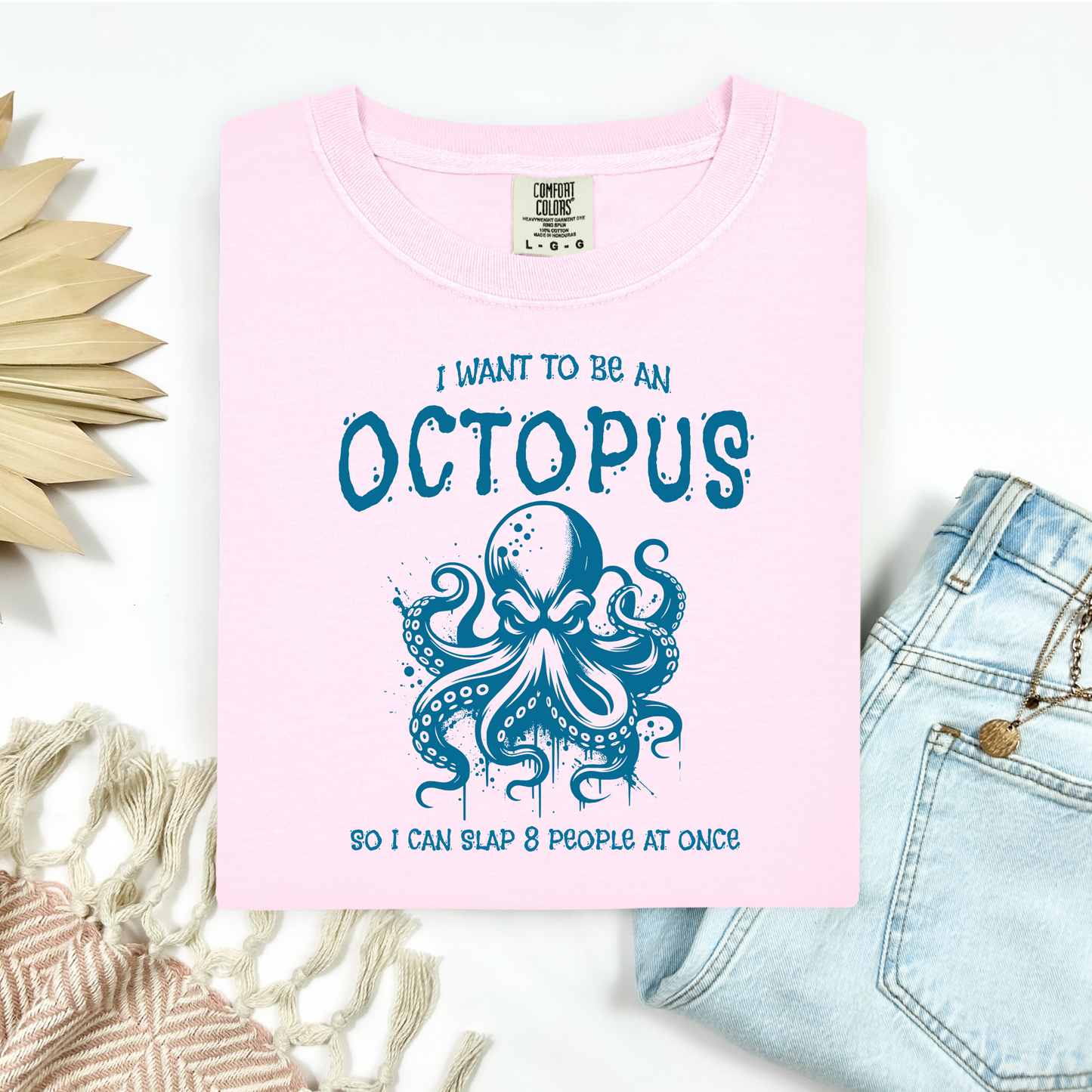 Comfort Colors 1717 "I Want To Be On Octopus So I Can Slap 8 People At Once" t shirt with an angry octopus one color graphic. Picture on a flay lay mockup with pair of blue jeans and blanket and a wicker sun shaped accessory.