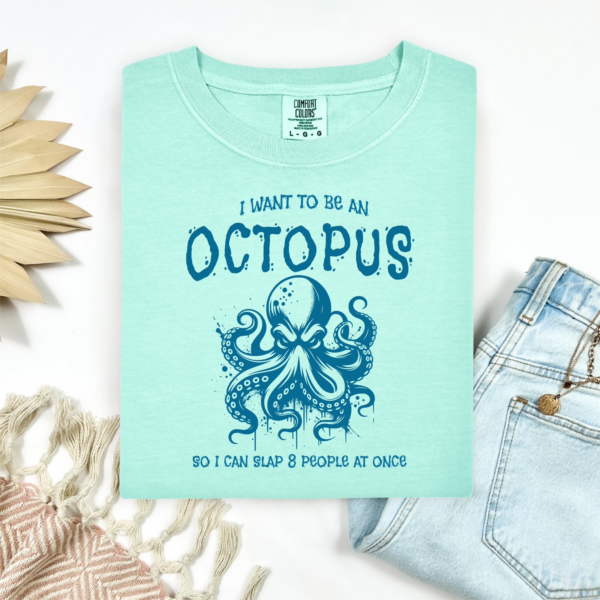 Comfort Colors 1717 "I Want To Be On Octopus So I Can Slap 8 People At Once" t shirt with an angry octopus one color graphic. Picture on a flay lay mockup with pair of blue jeans and blanket and a wicker sun shaped accessory.