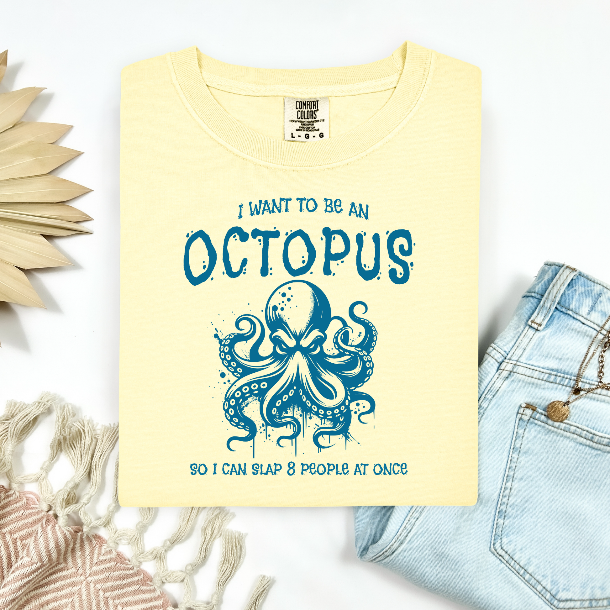 Comfort Colors 1717 "I Want To Be On Octopus So I Can Slap 8 People At Once" t shirt with an angry octopus one color graphic. Picture on a flay lay mockup with pair of blue jeans and blanket and a wicker sun shaped accessory.