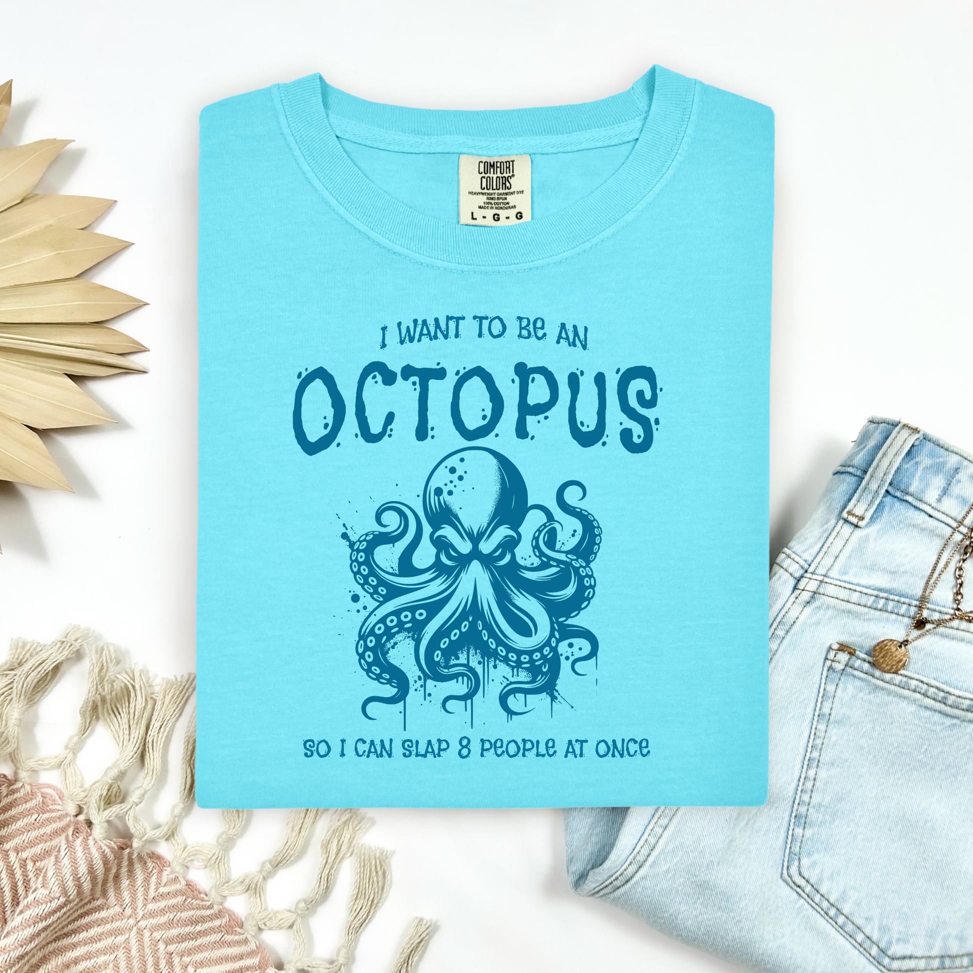 Comfort Colors 1717 "I Want To Be On Octopus So I Can Slap 8 People At Once" t shirt with an angry octopus one color graphic. Picture on a flay lay mockup with pair of blue jeans and blanket and a wicker sun shaped accessory.