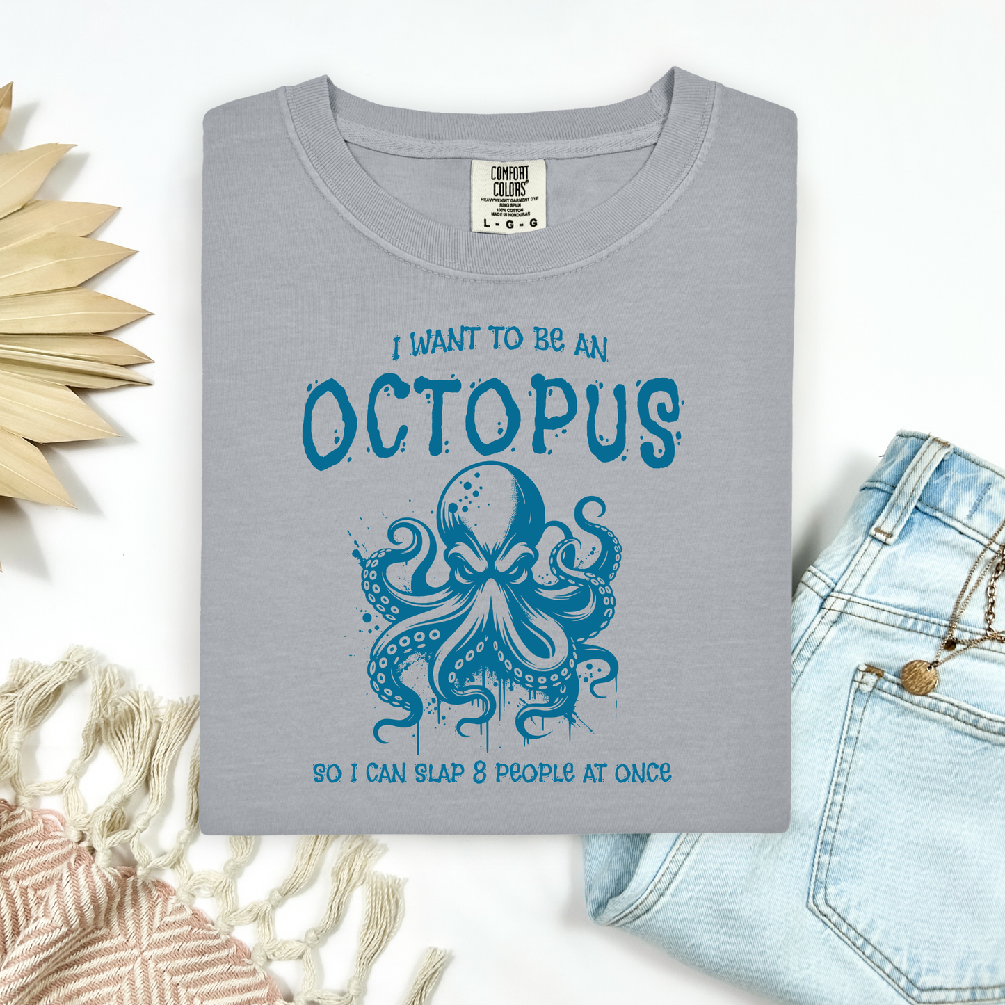 Comfort Colors 1717 "I Want To Be On Octopus So I Can Slap 8 People At Once" t shirt with an angry octopus one color graphic. Picture on a flay lay mockup with pair of blue jeans and blanket and a wicker sun shaped accessory.