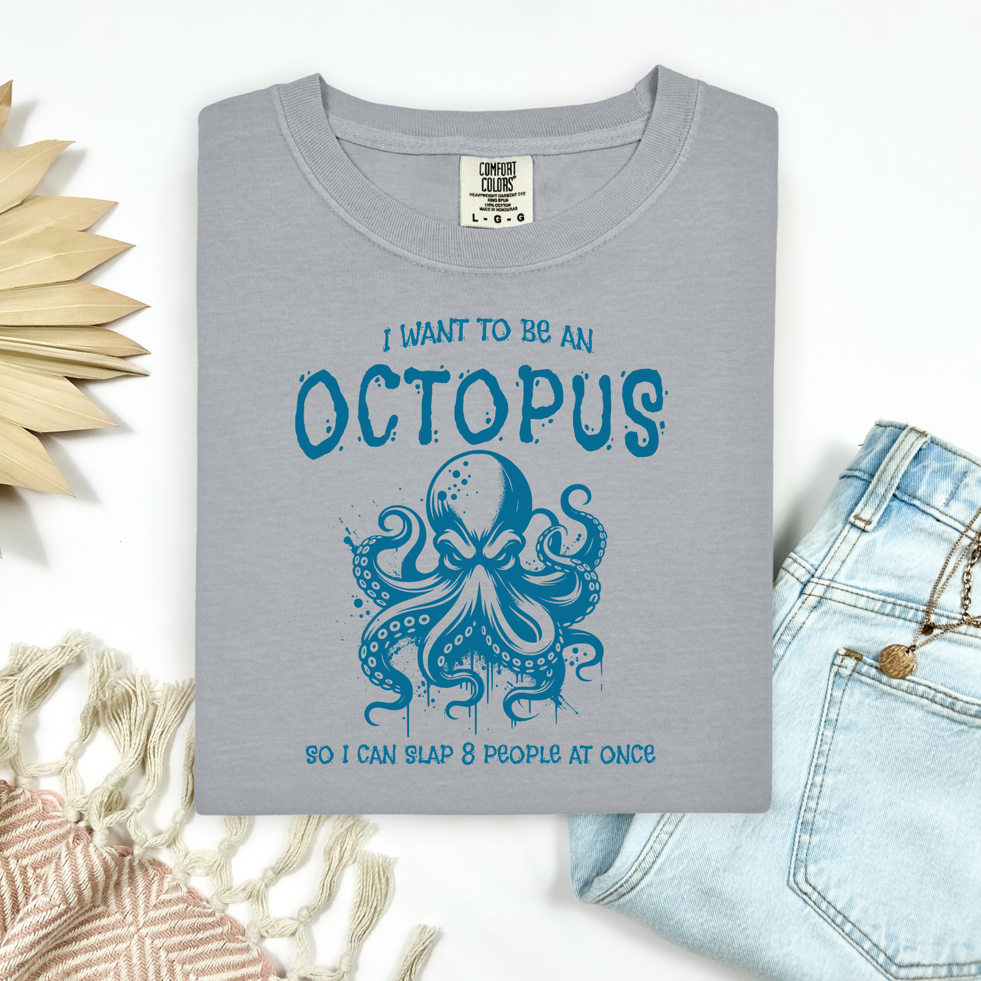 Comfort Colors 1717 "I Want To Be On Octopus So I Can Slap 8 People At Once" t shirt with an angry octopus one color graphic. Picture on a flay lay mockup with pair of blue jeans and blanket and a wicker sun shaped accessory.