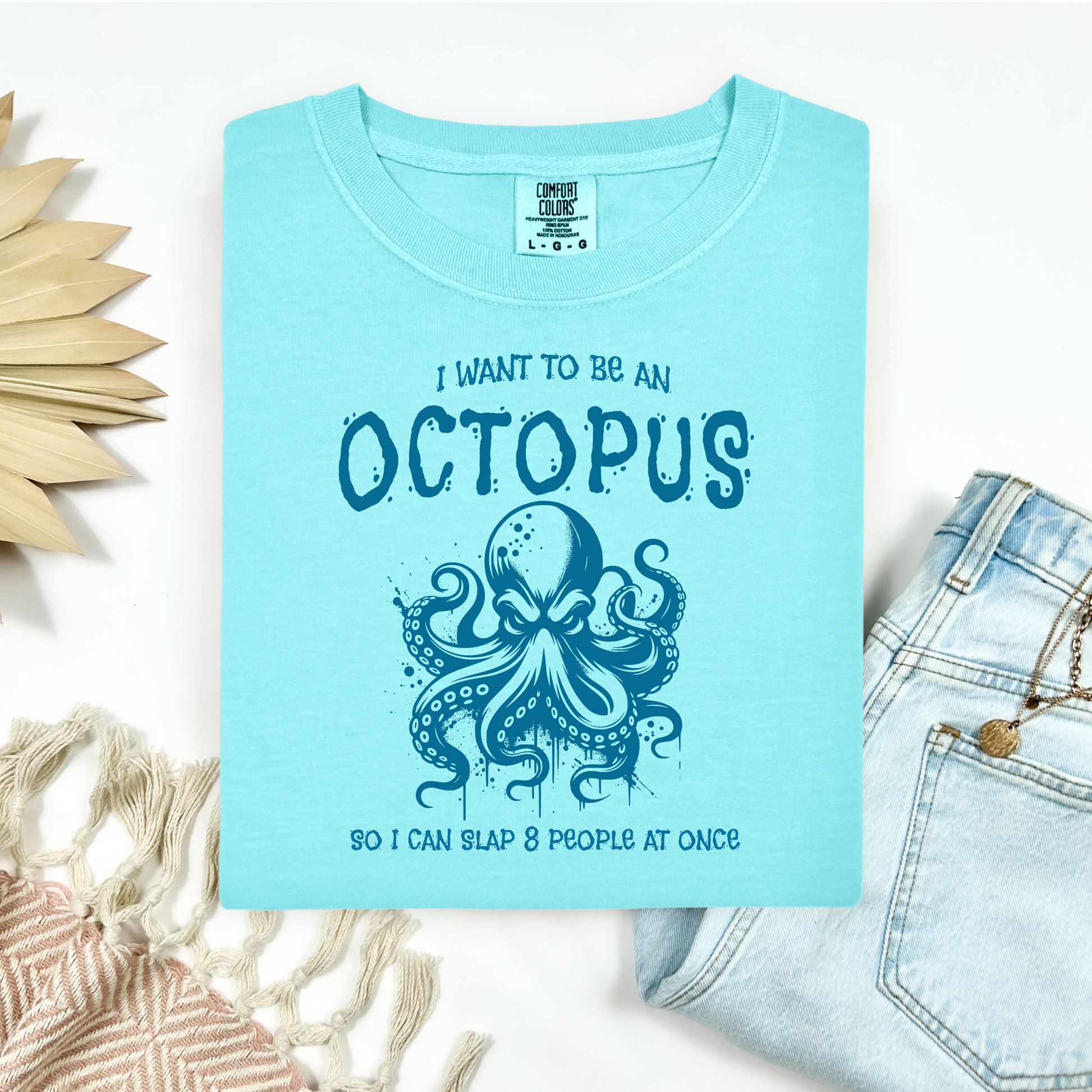 Comfort Colors 1717 "I Want To Be On Octopus So I Can Slap 8 People At Once" t shirt with an angry octopus one color graphic. Picture on a flay lay mockup with pair of blue jeans and blanket and a wicker sun shaped accessory.