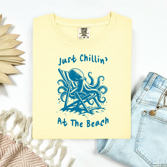 Comfort Colors 1717 "Just Chillin' At The Beach" t shirt with funny octopus lounging in a beach chair in a one color graphic. Picture on a flay lay mockup with pair of blue jeans and blanket and a wicker sun shaped accessory.