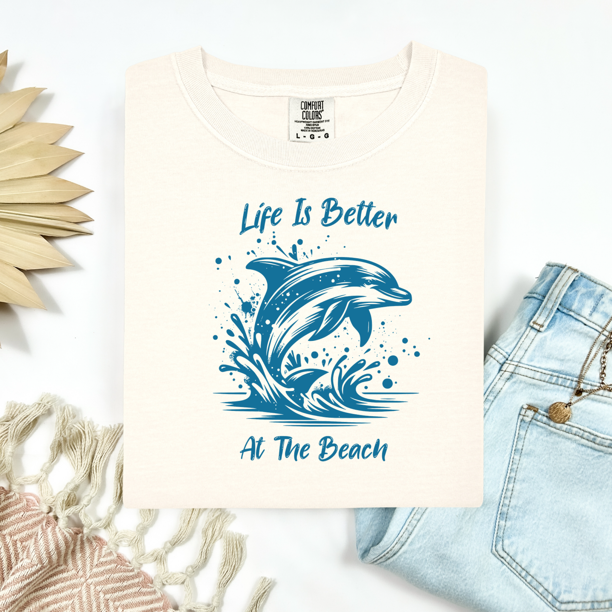 Comfort Colors 1717 "Life is Better at the Beach" t shirt with a dolphin jumping out of the water in one color graphic. Picture on a flay lay mockup with pair of blue jeans and blanket and a wicker sun shaped accessory.
