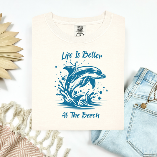 Comfort Colors 1717 "Life is Better at the Beach" t shirt with a dolphin jumping out of the water in one color graphic. Picture on a flay lay mockup with pair of blue jeans and blanket and a wicker sun shaped accessory.