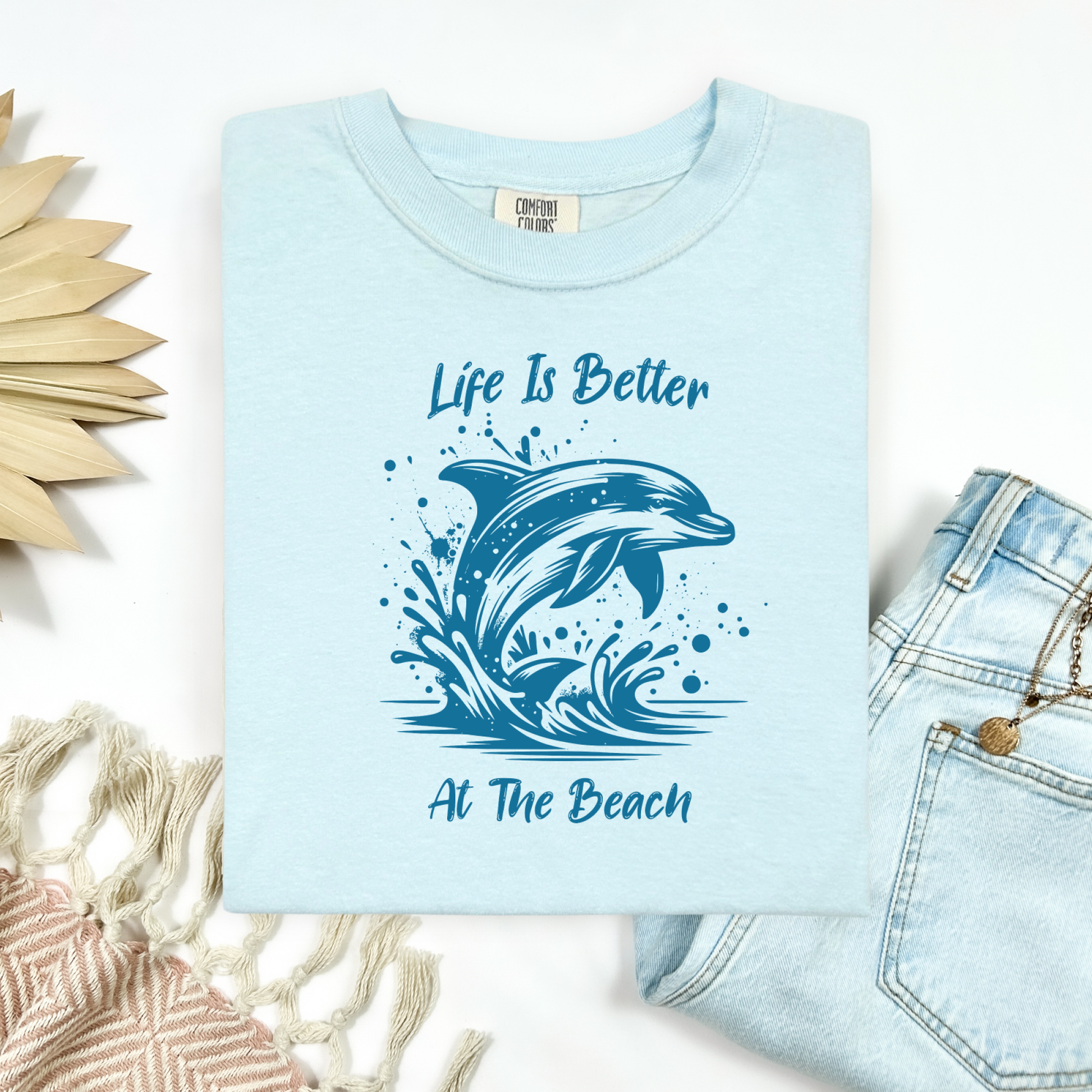 Comfort Colors 1717 "Life is Better at the Beach" t shirt with a dolphin jumping out of the water in one color graphic. Picture on a flay lay mockup with pair of blue jeans and blanket and a wicker sun shaped accessory.