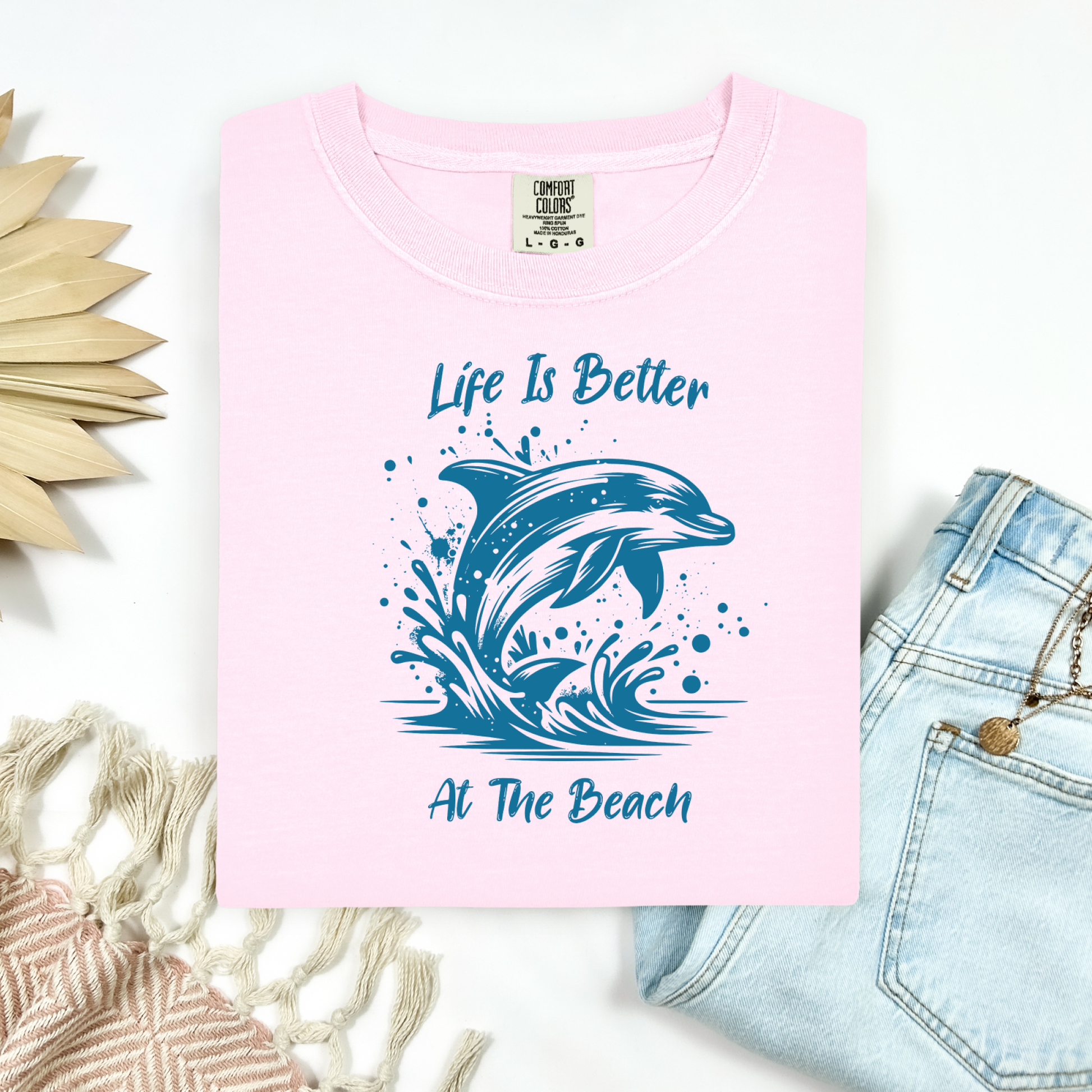 Comfort Colors 1717 "Life is Better at the Beach" t shirt with a dolphin jumping out of the water in one color graphic. Picture on a flay lay mockup with pair of blue jeans and blanket and a wicker sun shaped accessory.