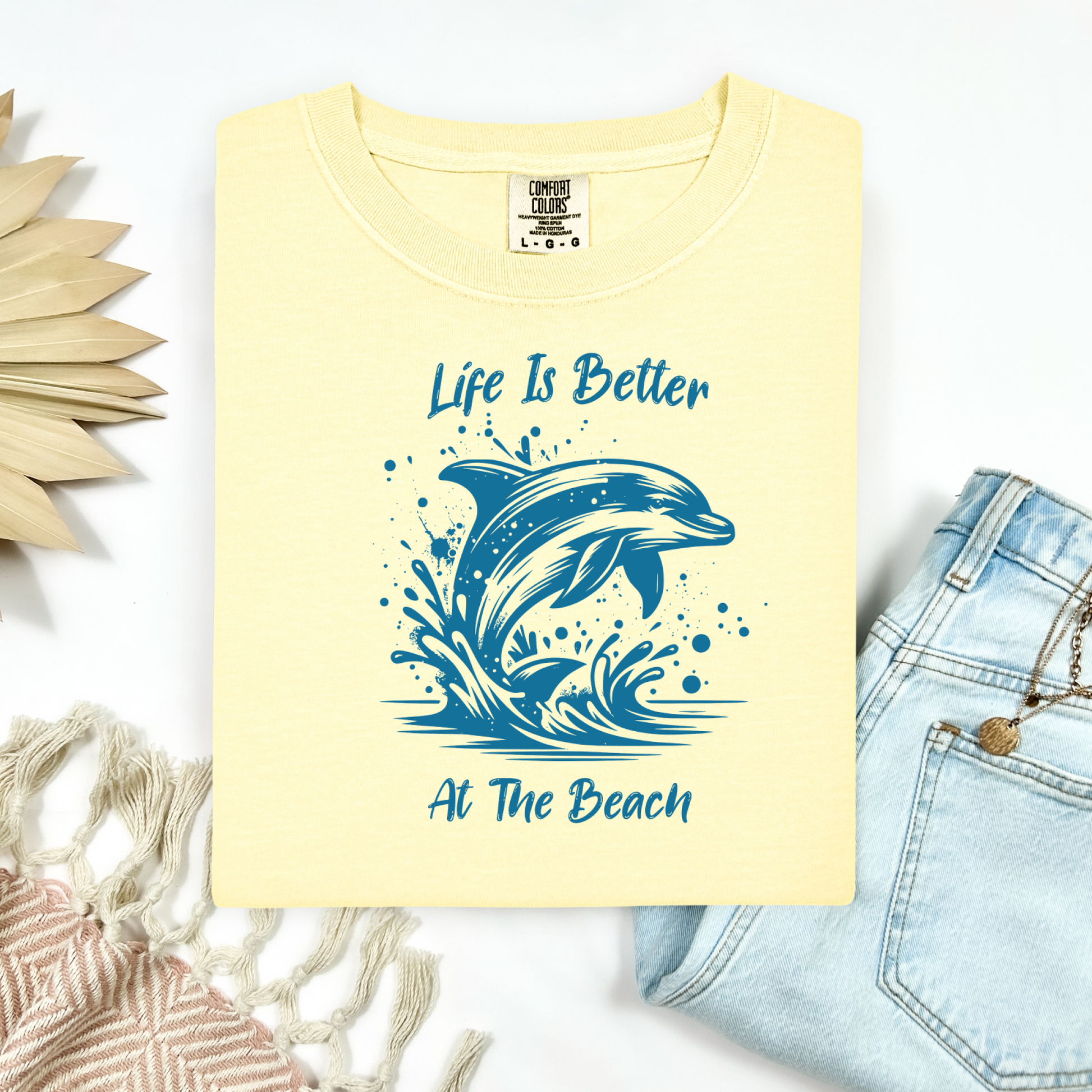 Comfort Colors 1717 "Life is Better at the Beach" t shirt with a dolphin jumping out of the water in one color graphic. Picture on a flay lay mockup with pair of blue jeans and blanket and a wicker sun shaped accessory.