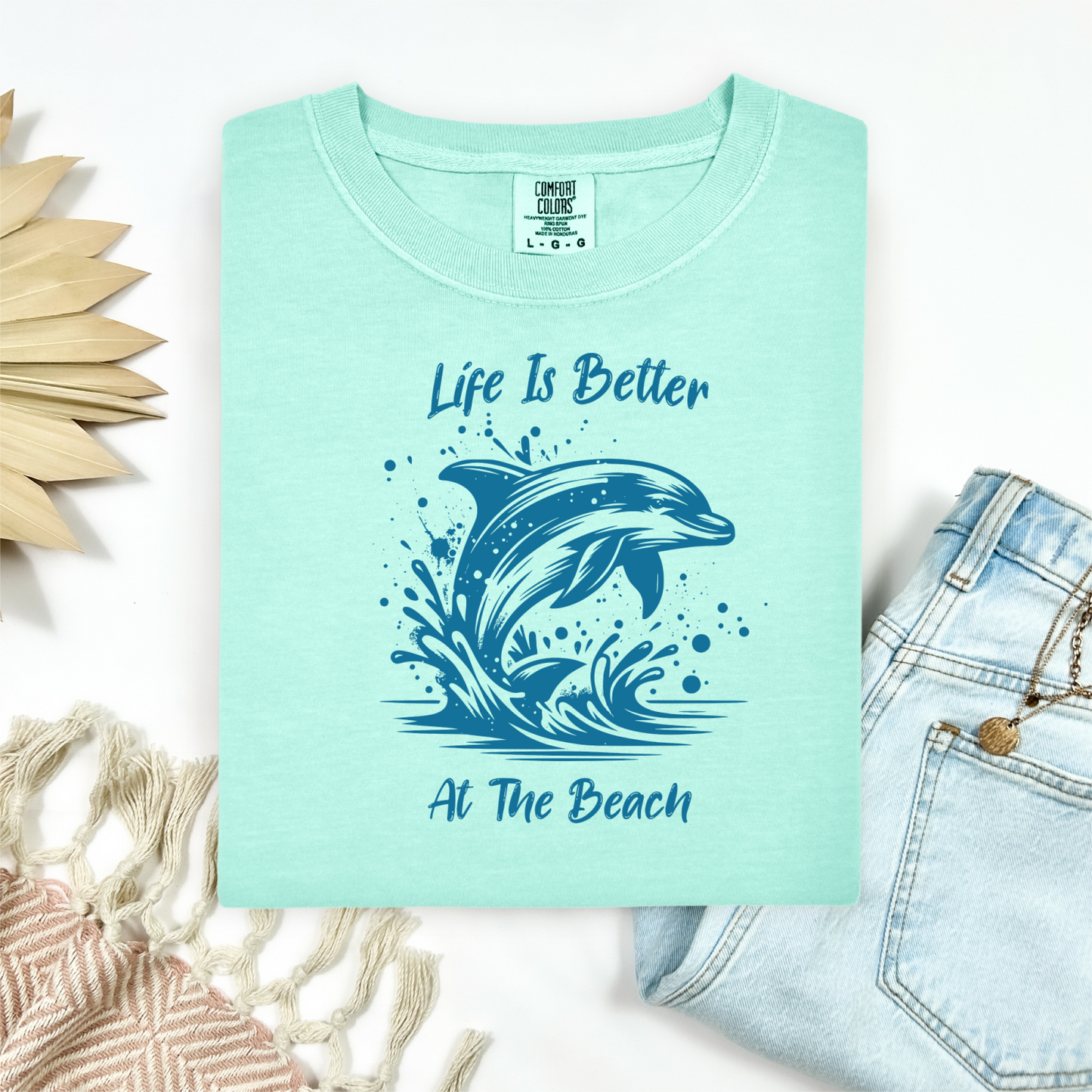 Comfort Colors 1717 "Life is Better at the Beach" t shirt with a dolphin jumping out of the water in one color graphic. Picture on a flay lay mockup with pair of blue jeans and blanket and a wicker sun shaped accessory.