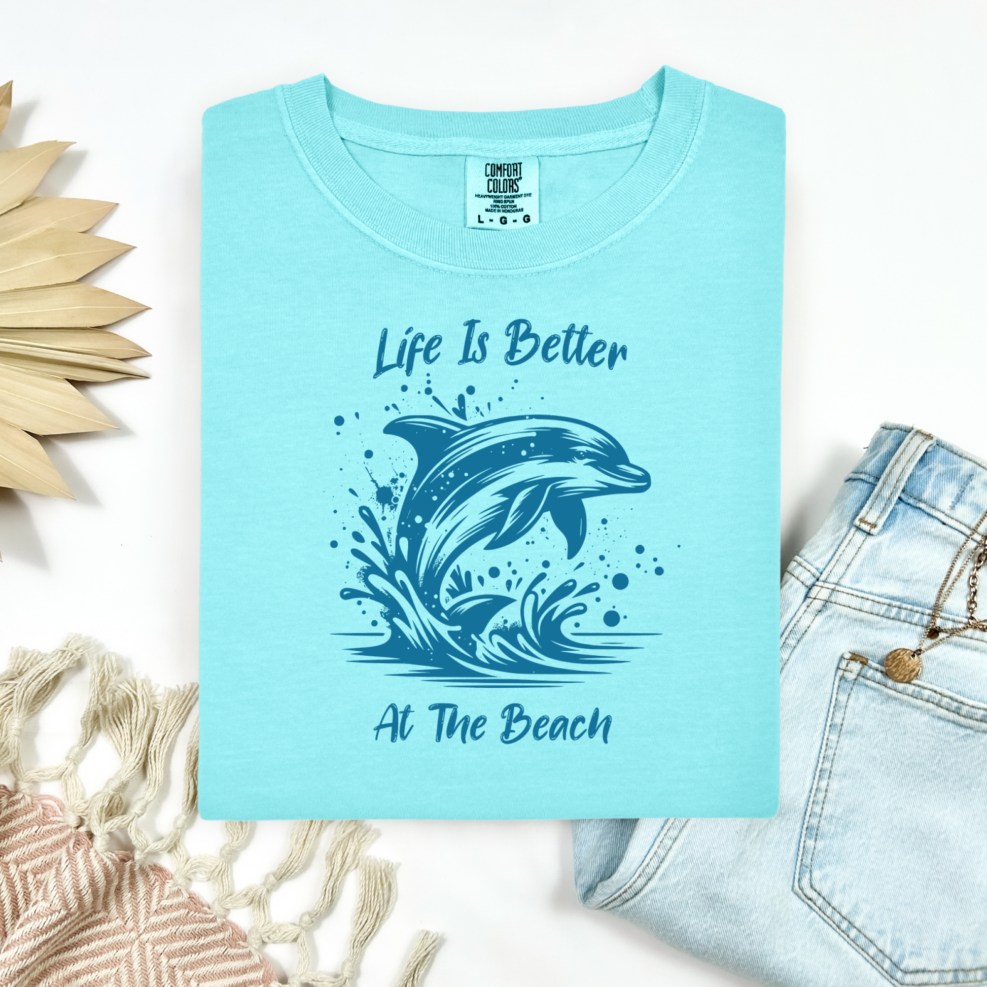 Comfort Colors 1717 "Life is Better at the Beach" t shirt with a dolphin jumping out of the water in one color graphic. Picture on a flay lay mockup with pair of blue jeans and blanket and a wicker sun shaped accessory.