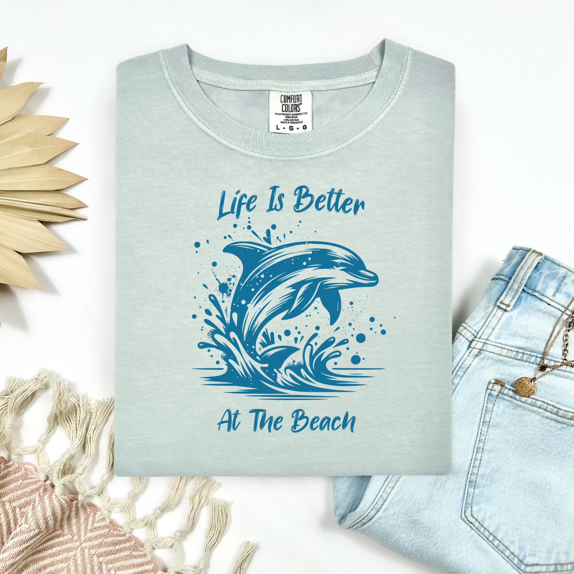 Comfort Colors 1717 "Life is Better at the Beach" t shirt with a dolphin jumping out of the water in one color graphic. Picture on a flay lay mockup with pair of blue jeans and blanket and a wicker sun shaped accessory.
