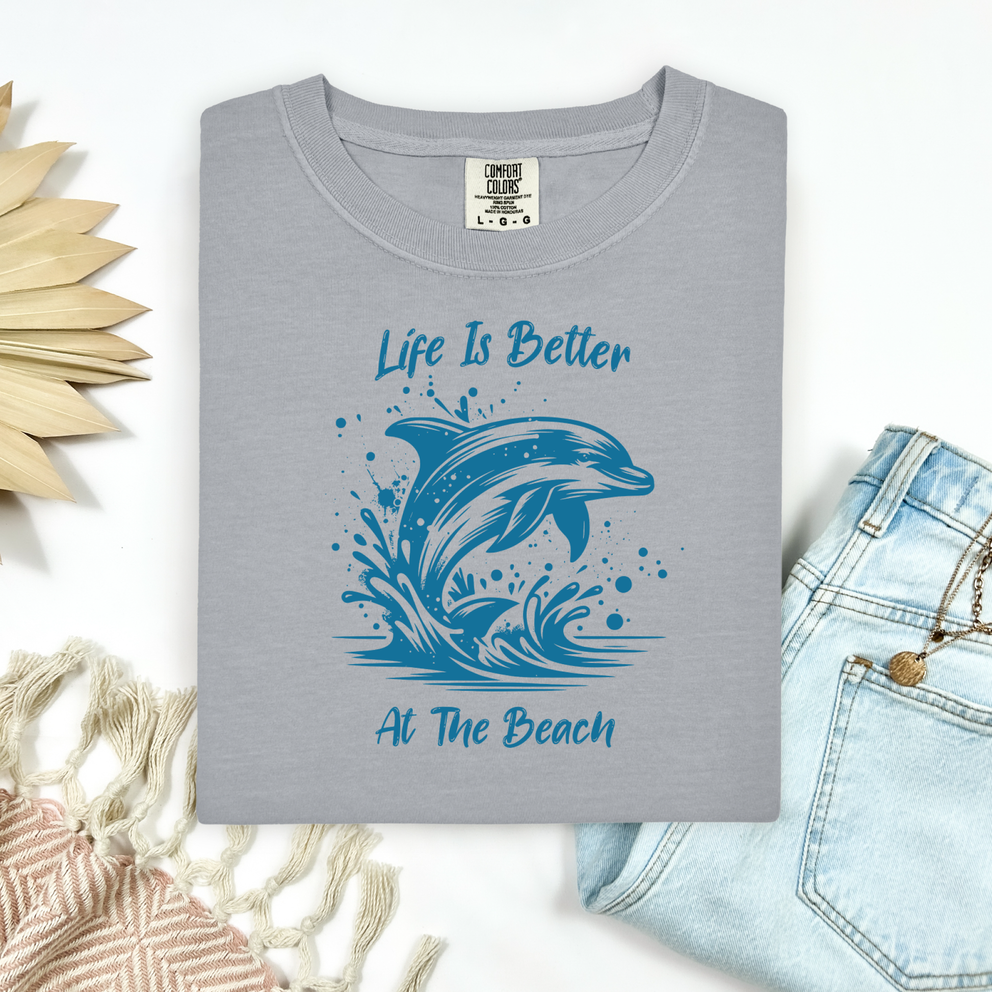 Comfort Colors 1717 "Life is Better at the Beach" t shirt with a dolphin jumping out of the water in one color graphic. Picture on a flay lay mockup with pair of blue jeans and blanket and a wicker sun shaped accessory.