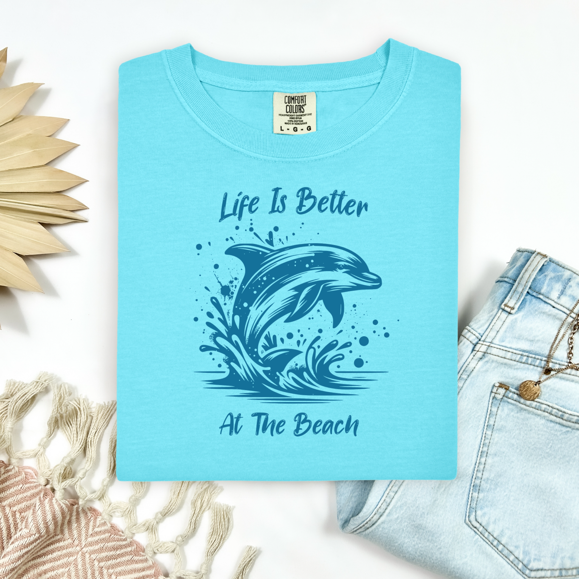 Comfort Colors 1717 "Life is Better at the Beach" t shirt with a dolphin jumping out of the water in one color graphic. Picture on a flay lay mockup with pair of blue jeans and blanket and a wicker sun shaped accessory.