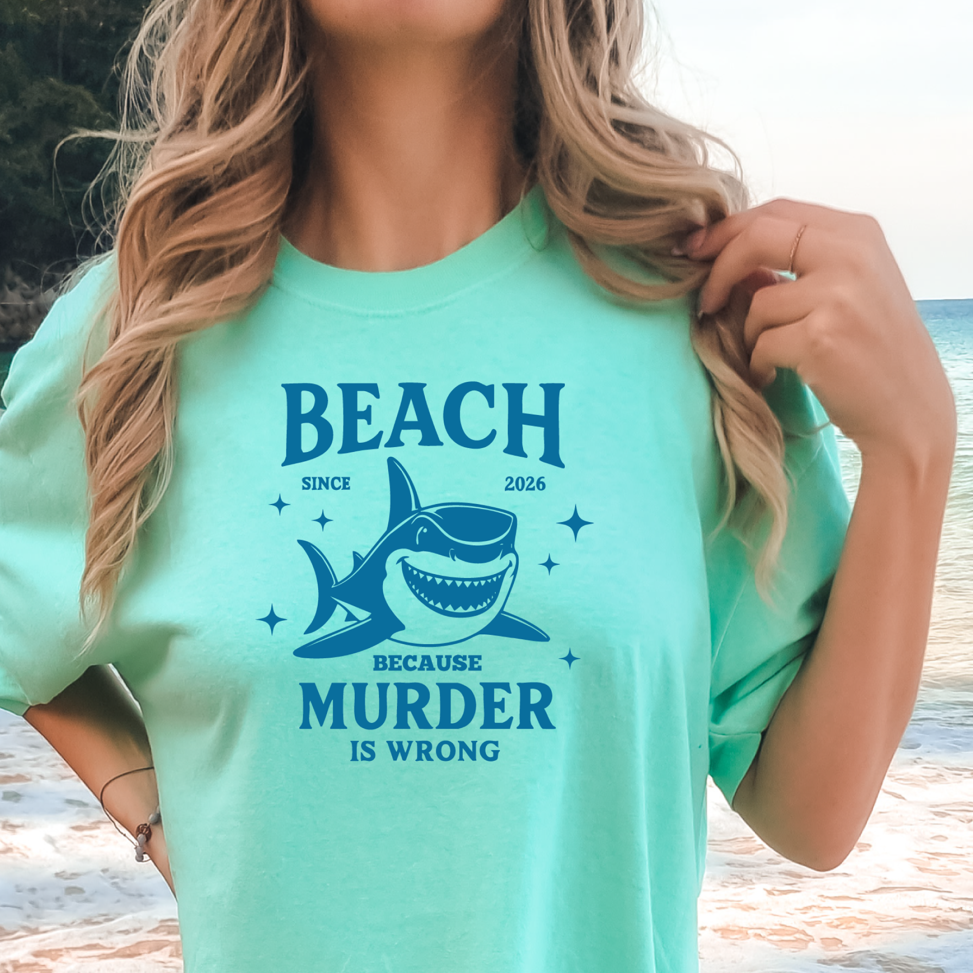 Comfort Colors t-shirt featuring a blue cartoon shark illustration with the humorous text “Beach Because Murder Is Wrong” and “Since 2026.” The design includes sparkling star accents for a fun coastal look. This unisex garment-dyed tee offers soft, vintage-style cotton and a relaxed fit, perfect for beach lovers, humor fans, and anyone who enjoys playful ocean-themed graphic shirts.