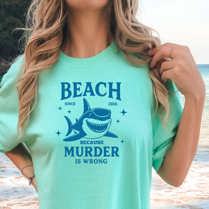 Comfort Colors t-shirt featuring a blue cartoon shark illustration with the humorous text “Beach Because Murder Is Wrong” and “Since 2026.” The design includes sparkling star accents for a fun coastal look. This unisex garment-dyed tee offers soft, vintage-style cotton and a relaxed fit, perfect for beach lovers, humor fans, and anyone who enjoys playful ocean-themed graphic shirts.
