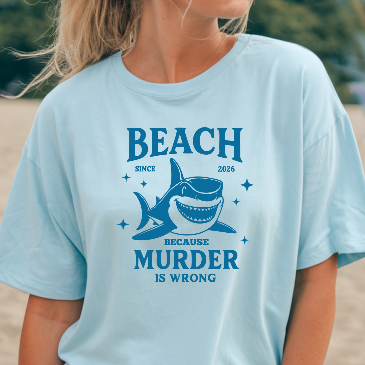 Comfort Colors t-shirt featuring a blue cartoon shark illustration with the humorous text “Beach Because Murder Is Wrong” and “Since 2026.” The design includes sparkling star accents for a fun coastal look. This unisex garment-dyed tee offers soft, vintage-style cotton and a relaxed fit, perfect for beach lovers, humor fans, and anyone who enjoys playful ocean-themed graphic shirts.