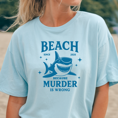 Comfort Colors t-shirt featuring a blue cartoon shark illustration with the humorous text “Beach Because Murder Is Wrong” and “Since 2026.” The design includes sparkling star accents for a fun coastal look. This unisex garment-dyed tee offers soft, vintage-style cotton and a relaxed fit, perfect for beach lovers, humor fans, and anyone who enjoys playful ocean-themed graphic shirts.