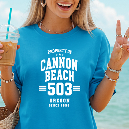 Woman wearing a blue t-shirt with 'Cannon Beach 503' text, holding a drink by the beach.