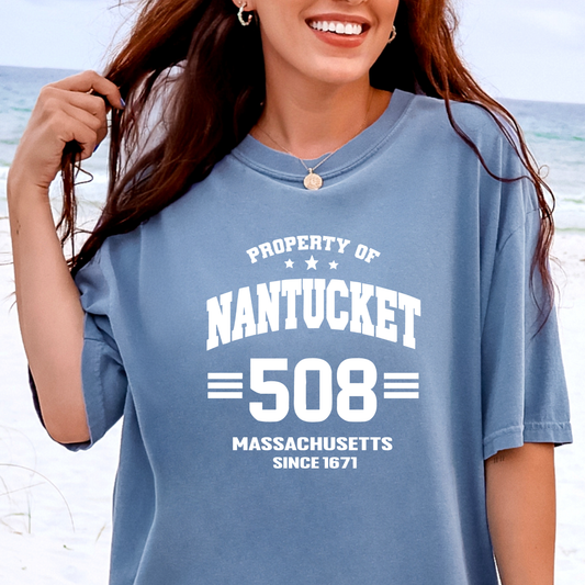 Person wearing a blue t-shirt with 'Property of Nantucket 508 Massachusetts Since 1671' text on a beach.