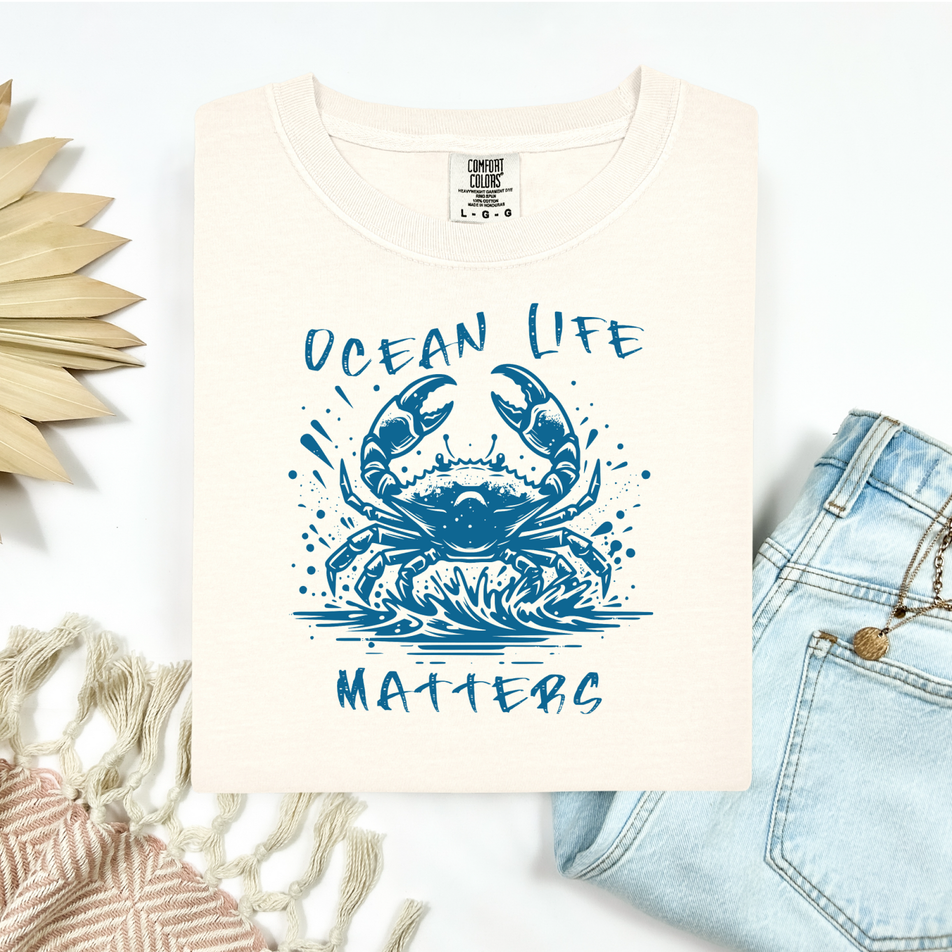 Comfort Colors 1717 "Ocean Life Matters" t shirt with a crab splashing in water in a one color graphic. Picture on a flay lay mockup with pair of blue jeans and blanket and a wicker sun shaped accessory.