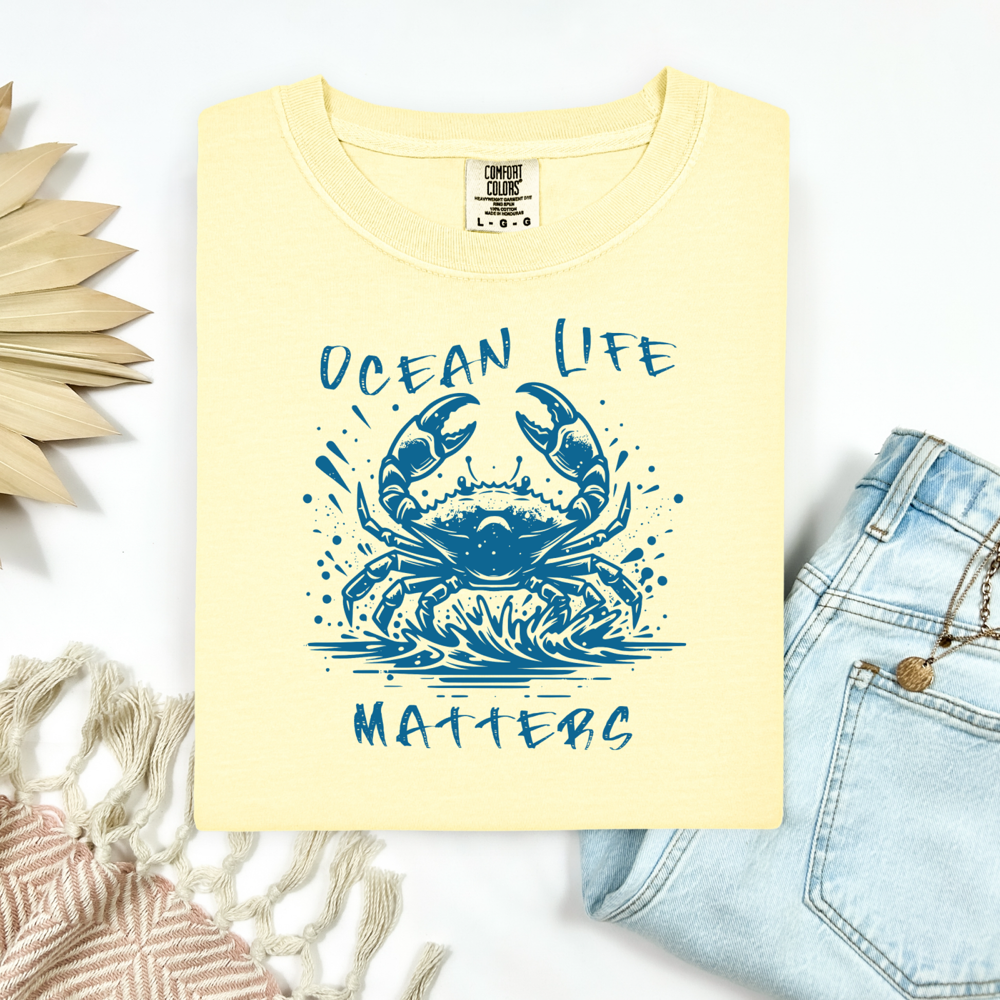 Comfort Colors 1717 "Ocean Life Matters" t shirt with a crab splashing in water in a one color graphic. Picture on a flay lay mockup with pair of blue jeans and blanket and a wicker sun shaped accessory.
