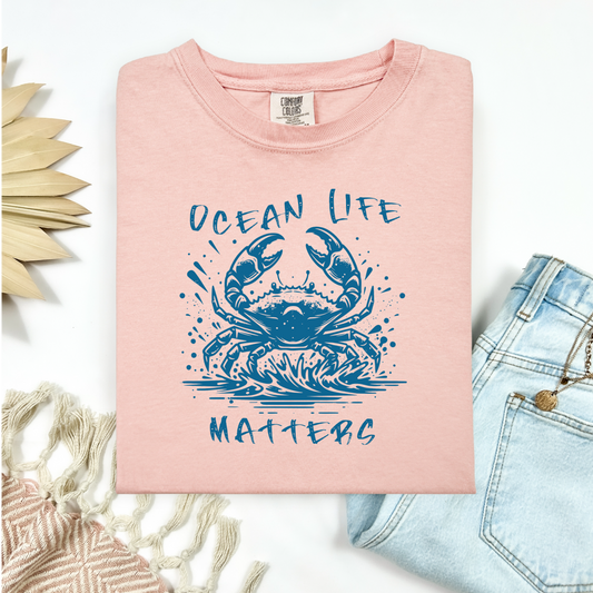 Comfort Colors 1717 "Ocean Life Matters" t shirt with a crab splashing in water in a one color graphic. Picture on a flay lay mockup with pair of blue jeans and blanket and a wicker sun shaped accessory.