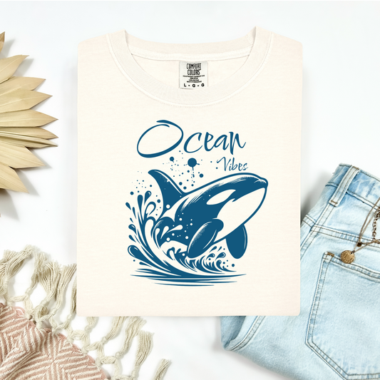 Comfort Colors 1717 "Ocean Vibes" t shirt with a orca whale jumping out of the water in one color graphic. Picture on a flay lay mockup with pair of blue jeans and blanket and a wicker sun shaped accessory.