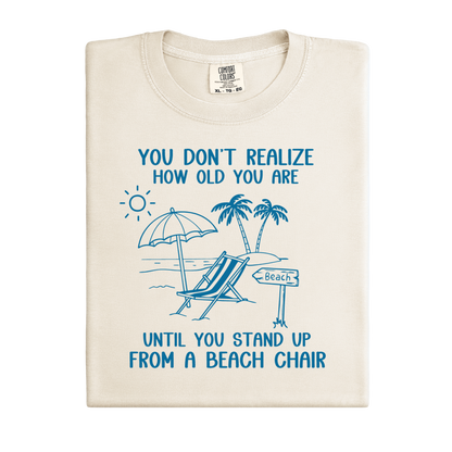 Beige t-shirt with blue beach-themed design and text on a white background