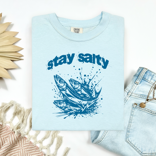 Comfort Colors 1717 "Stay Salty" t shirt with three sardines splashing in water in a one color graphic. Picture on a flay lay mockup with pair of blue jeans and blanket and a wicker sun shaped accessory.