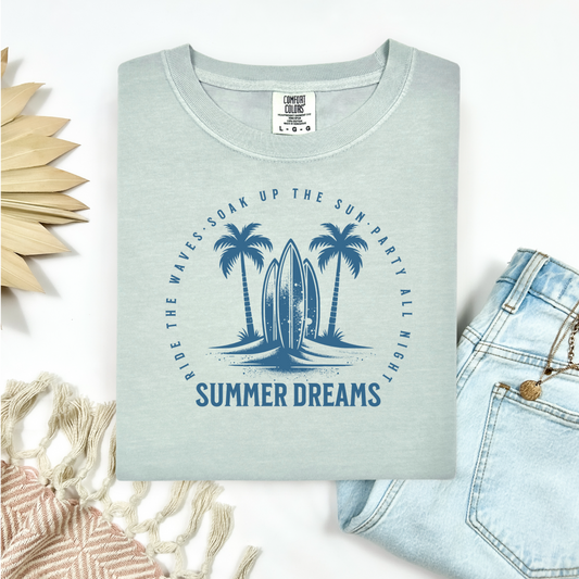 Comfort Colors 1717 "Summer Dreams" t shirt with surfboard and palm trees surrounded by inspiring text in one color graphic. Picture on a flay lay mockup with pair of blue jeans and blanket and a wicker sun shaped accessory.
