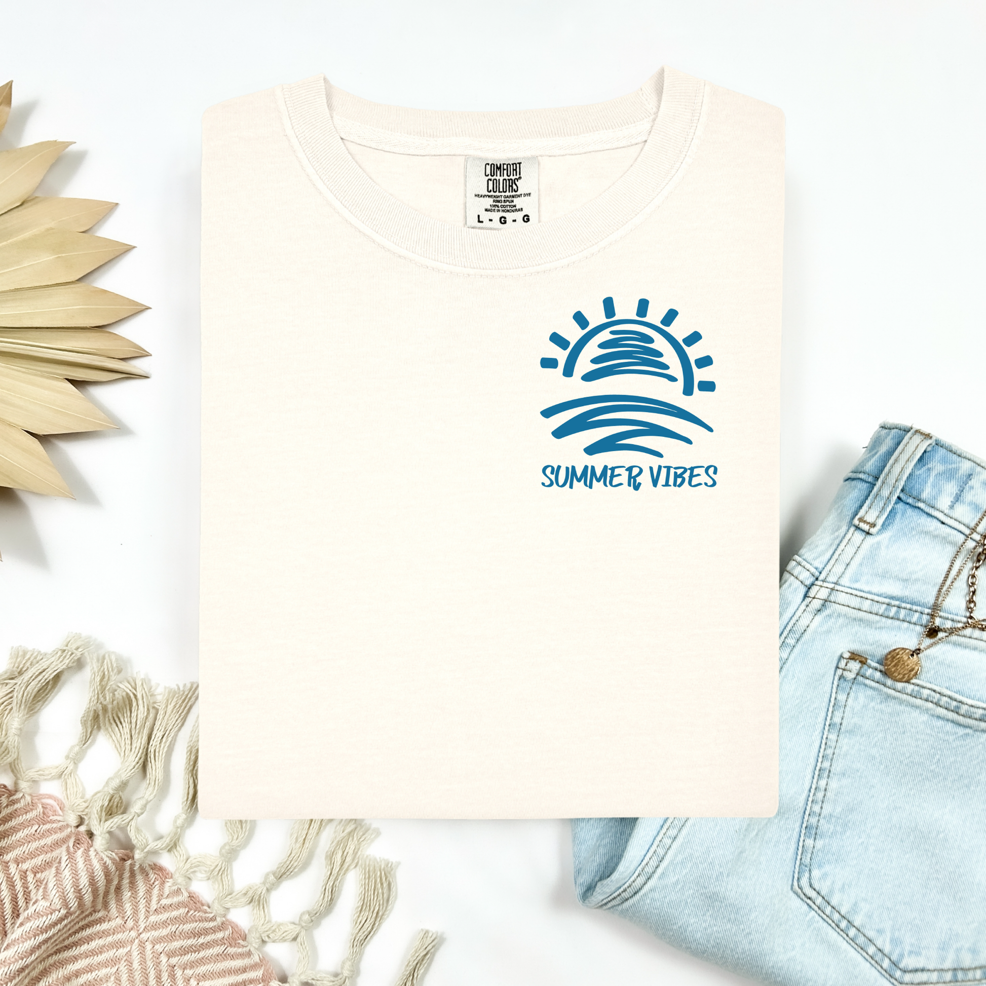 Comfort Colors 1717 "Summer Vibes" t shirt with sun and waves one color graphic. Picture on a flay lay mockup with pair of blue jeans and blanket and a wicker sun shaped accessory.