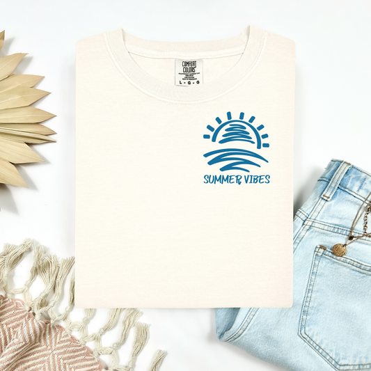 Comfort Colors 1717 "Summer Vibes" t shirt with sun and waves one color graphic. Picture on a flay lay mockup with pair of blue jeans and blanket and a wicker sun shaped accessory.