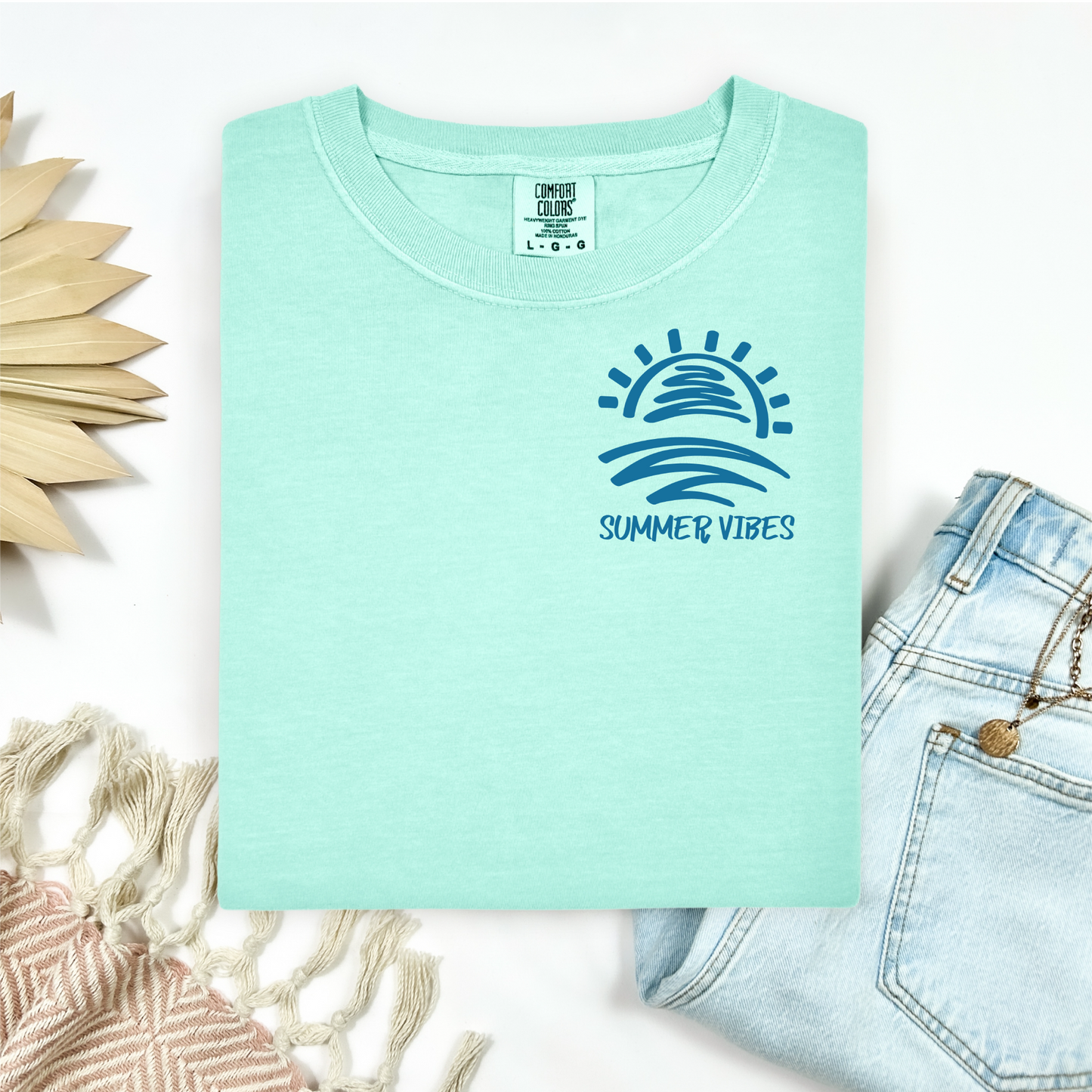 Comfort Colors 1717 "Summer Vibes" t shirt with sun and waves one color graphic. Picture on a flay lay mockup with pair of blue jeans and blanket and a wicker sun shaped accessory.