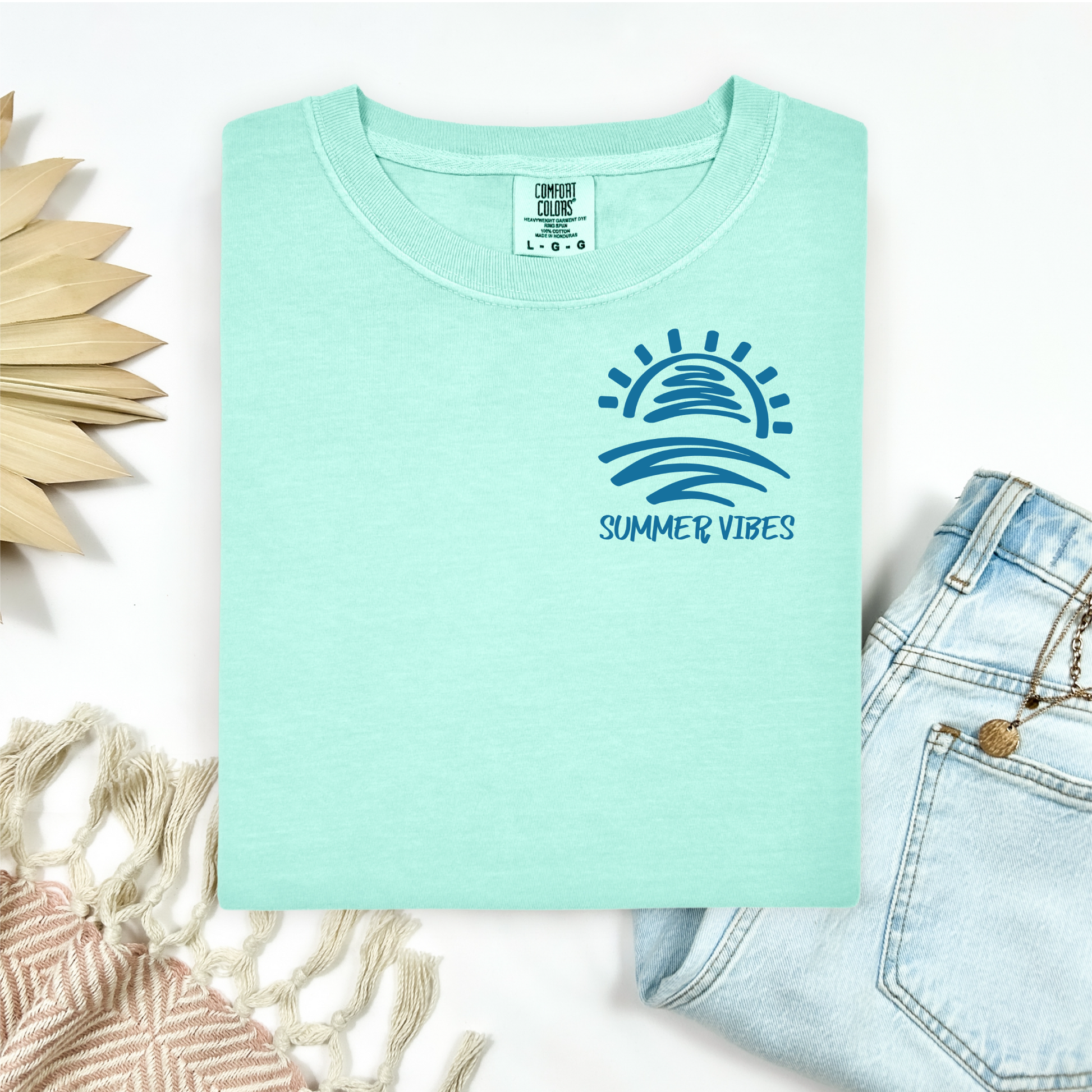 Comfort Colors 1717 "Summer Vibes" t shirt with sun and waves one color graphic. Picture on a flay lay mockup with pair of blue jeans and blanket and a wicker sun shaped accessory.