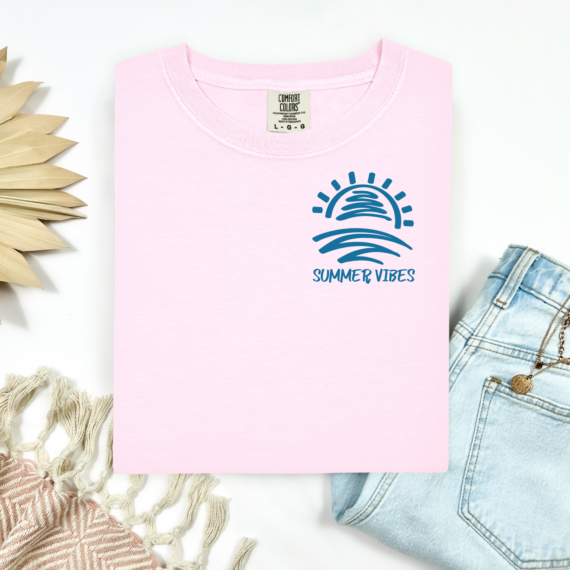 Comfort Colors 1717 "Summer Vibes" t shirt with sun and waves one color graphic. Picture on a flay lay mockup with pair of blue jeans and blanket and a wicker sun shaped accessory.