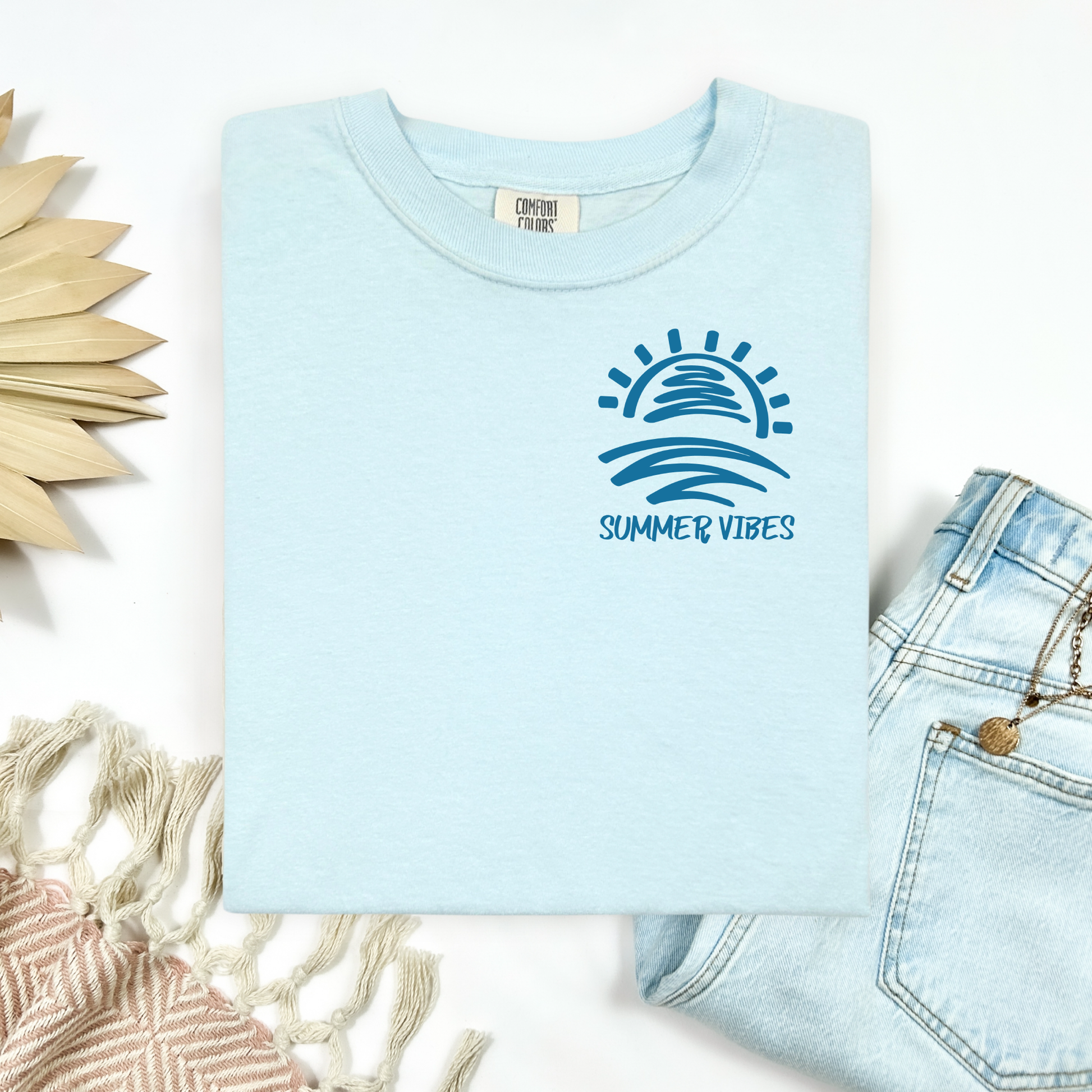 Comfort Colors 1717 "Summer Vibes" t shirt with sun and waves one color graphic. Picture on a flay lay mockup with pair of blue jeans and blanket and a wicker sun shaped accessory.