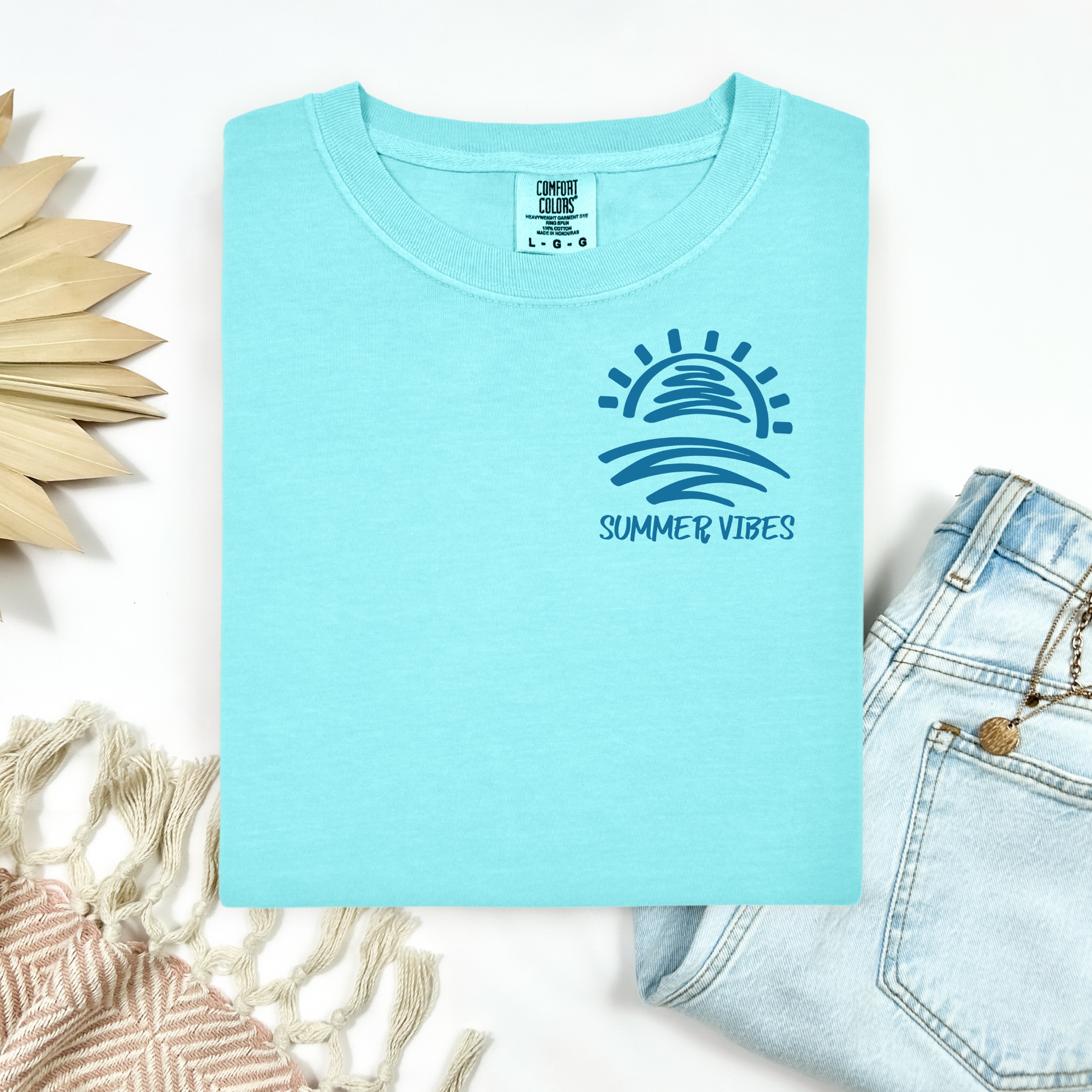 Comfort Colors 1717 "Summer Vibes" t shirt with sun and waves one color graphic. Picture on a flay lay mockup with pair of blue jeans and blanket and a wicker sun shaped accessory.