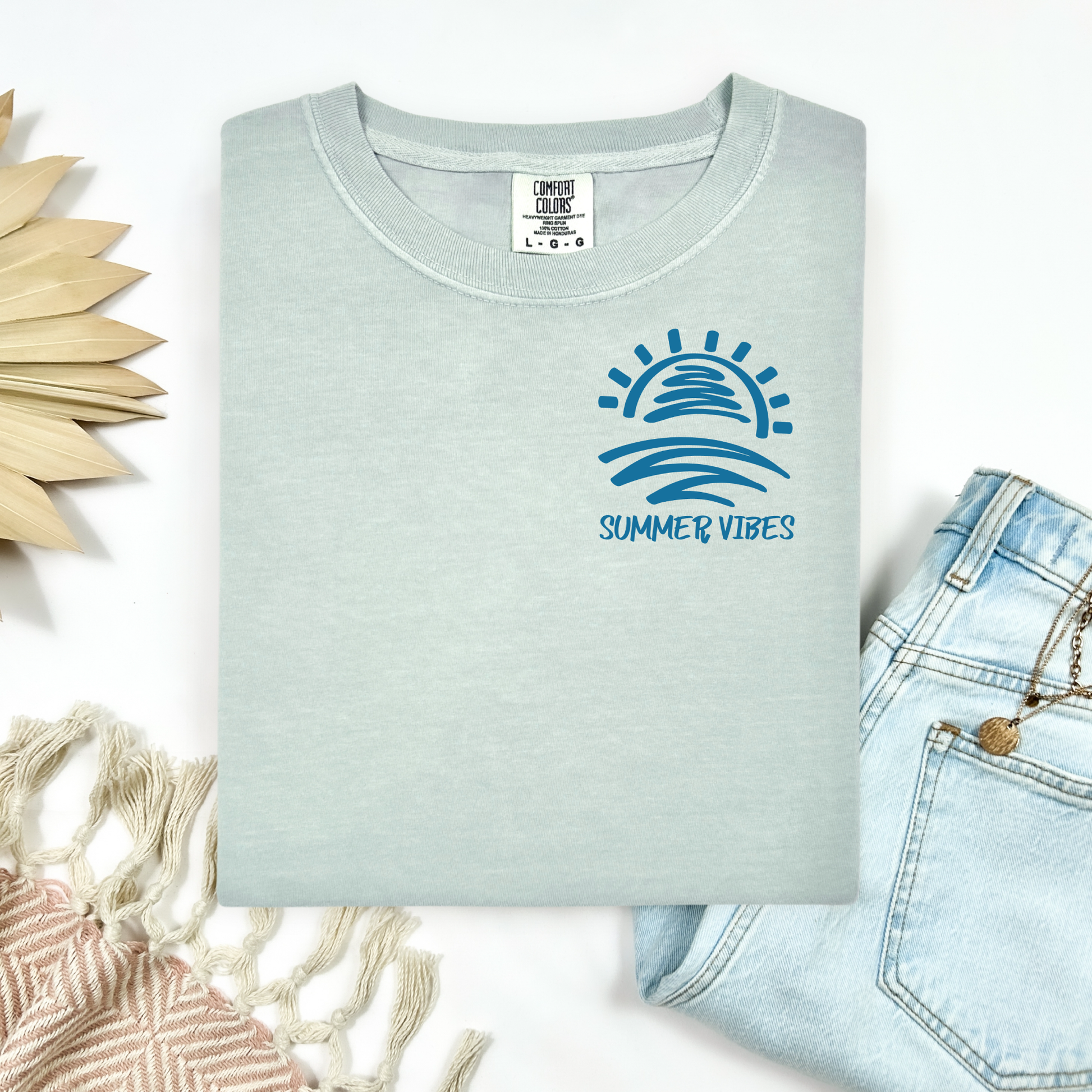 Comfort Colors 1717 "Summer Vibes" t shirt with sun and waves one color graphic. Picture on a flay lay mockup with pair of blue jeans and blanket and a wicker sun shaped accessory.