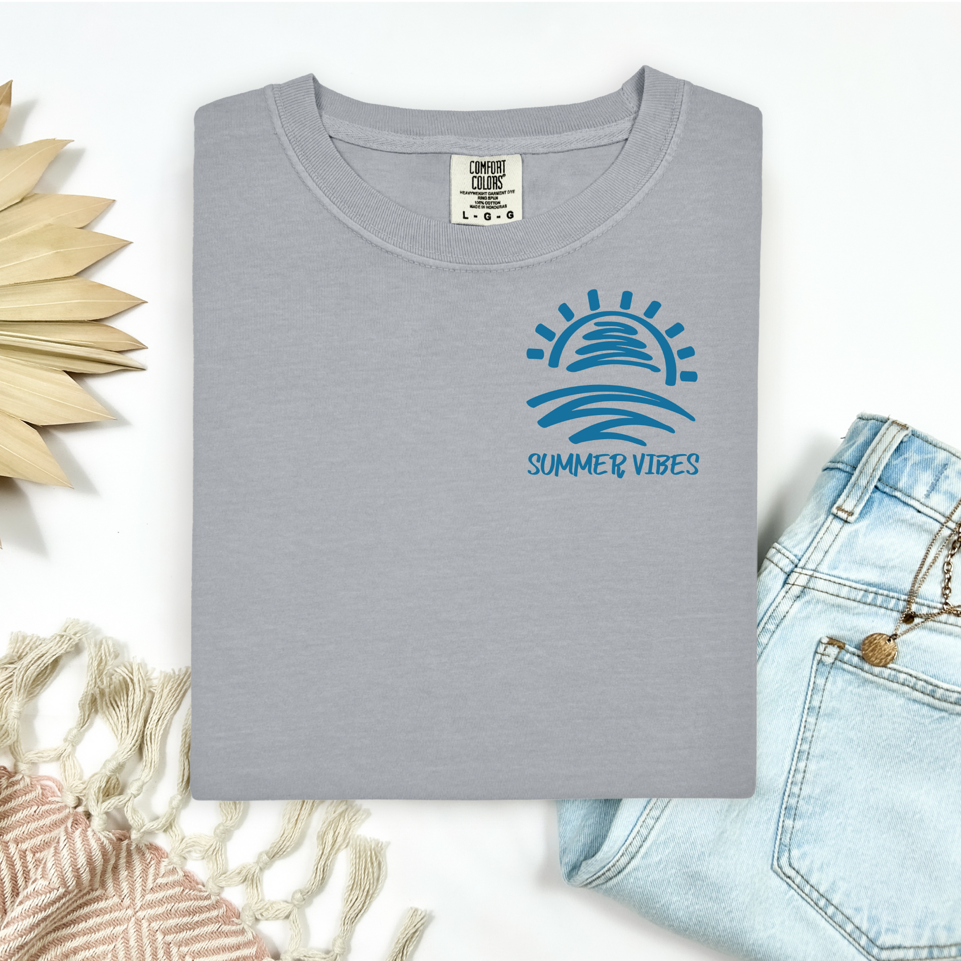 Comfort Colors 1717 "Summer Vibes" t shirt with sun and waves one color graphic. Picture on a flay lay mockup with pair of blue jeans and blanket and a wicker sun shaped accessory.