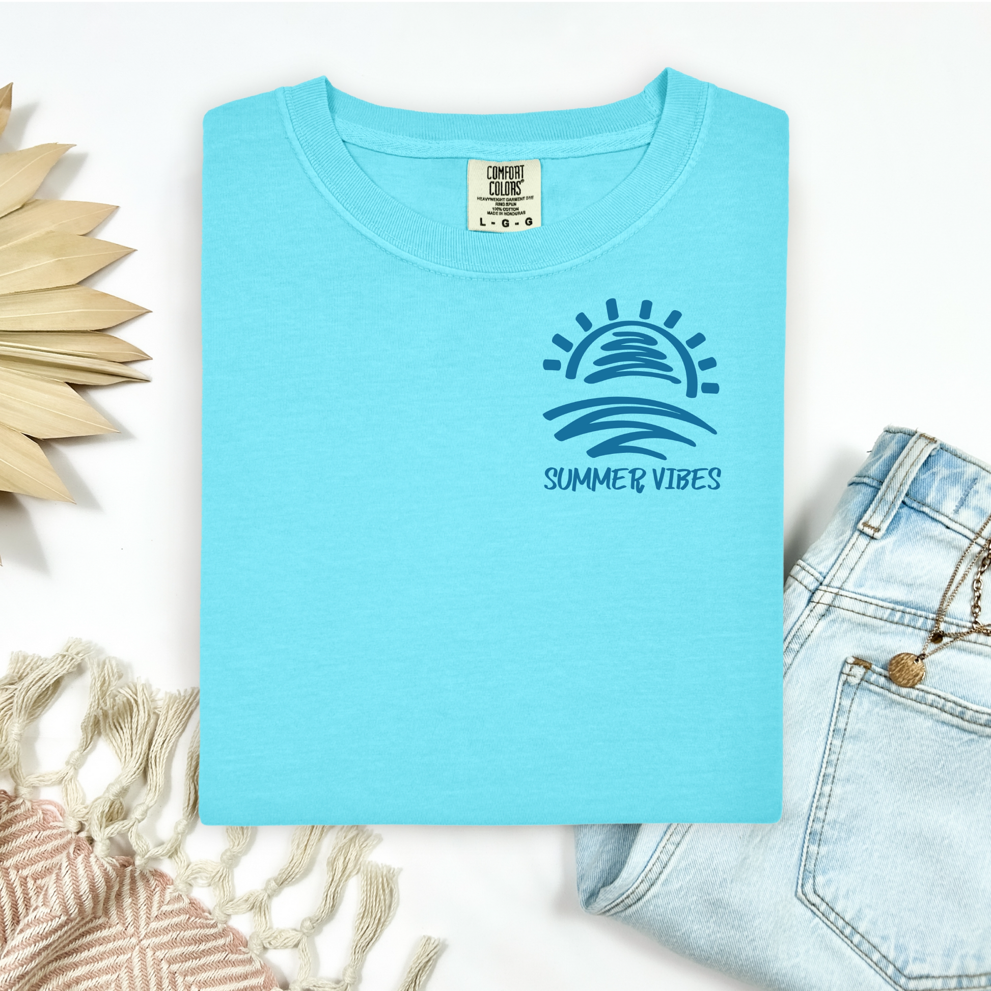 Comfort Colors 1717 "Summer Vibes" t shirt with sun and waves one color graphic. Picture on a flay lay mockup with pair of blue jeans and blanket and a wicker sun shaped accessory.