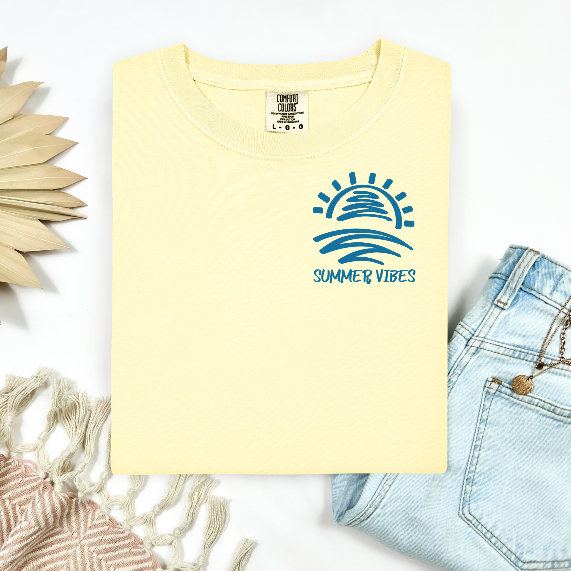 Comfort Colors 1717 "Summer Vibes" t shirt with sun and waves one color graphic. Picture on a flay lay mockup with pair of blue jeans and blanket and a wicker sun shaped accessory.