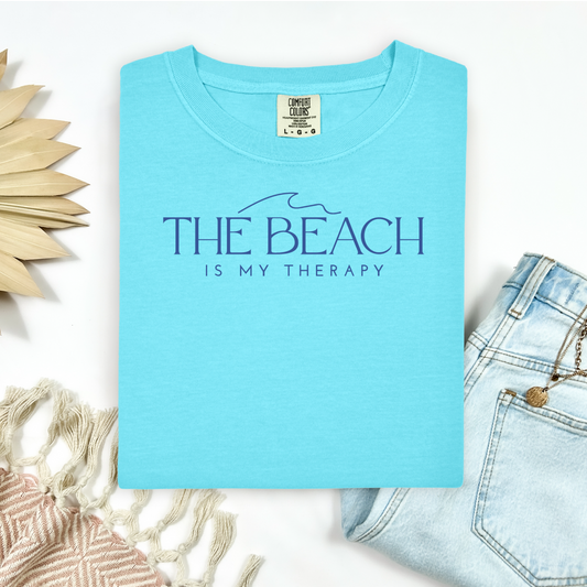 Comfort Colors 1717 "The Beach Is My Therapy" t shirt with a simple wave and one color graphic. Picture on a flay lay mockup with pair of blue jeans and blanket and a wicker sun shaped accessory.