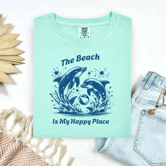 Comfort Colors 1717 "The Beach Is My Happy Place" t shirt with playful dolphins with a beach ball in one color graphic. Picture on a flay lay mockup with pair of blue jeans and blanket and a wicker sun shaped accessory.