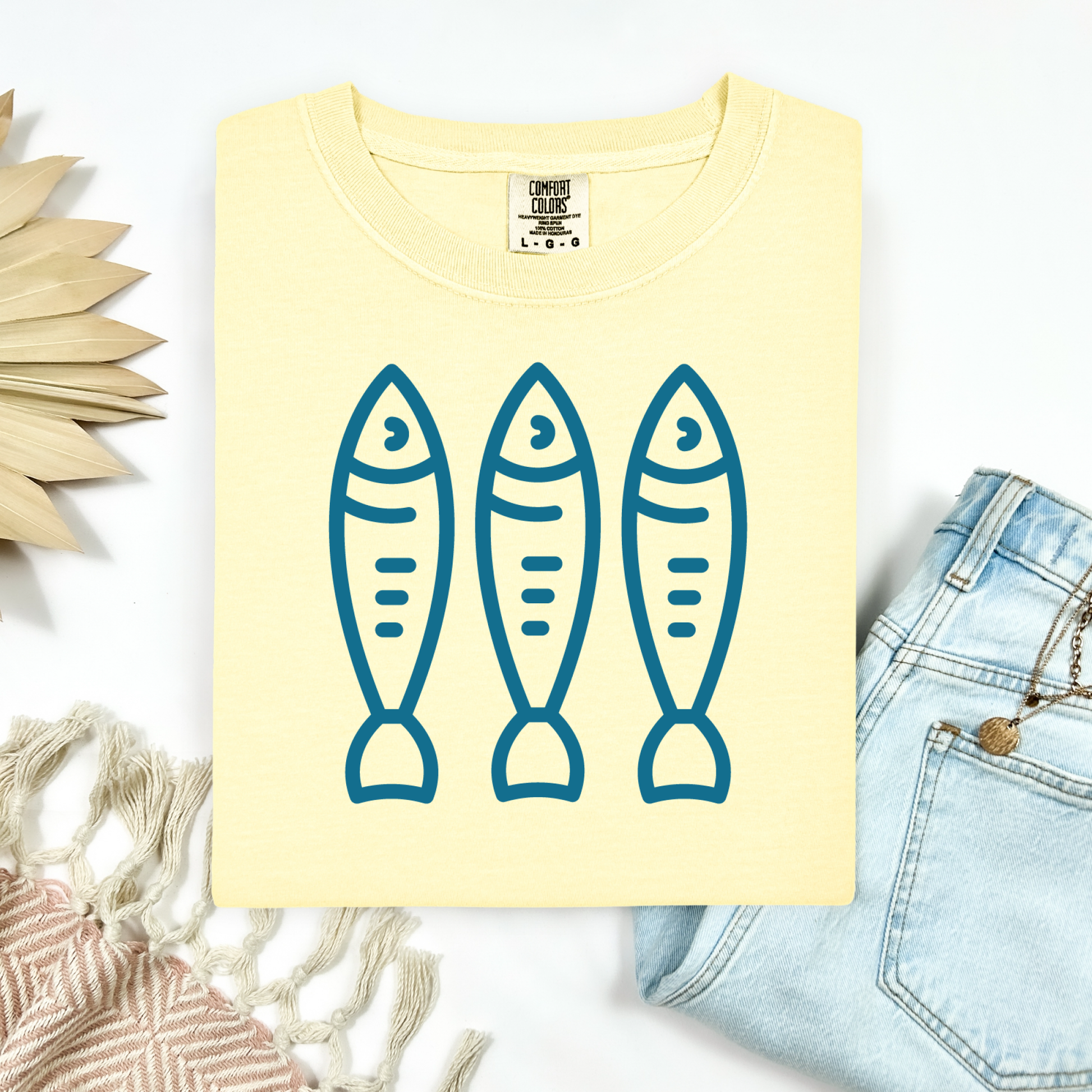 Comfort Colors 1717 Fisherman's Aesthetic t shirt with three sardines in a  one color graphic. Picture on a flay lay mockup with pair of blue jeans and blanket and a wicker sun shaped accessory.