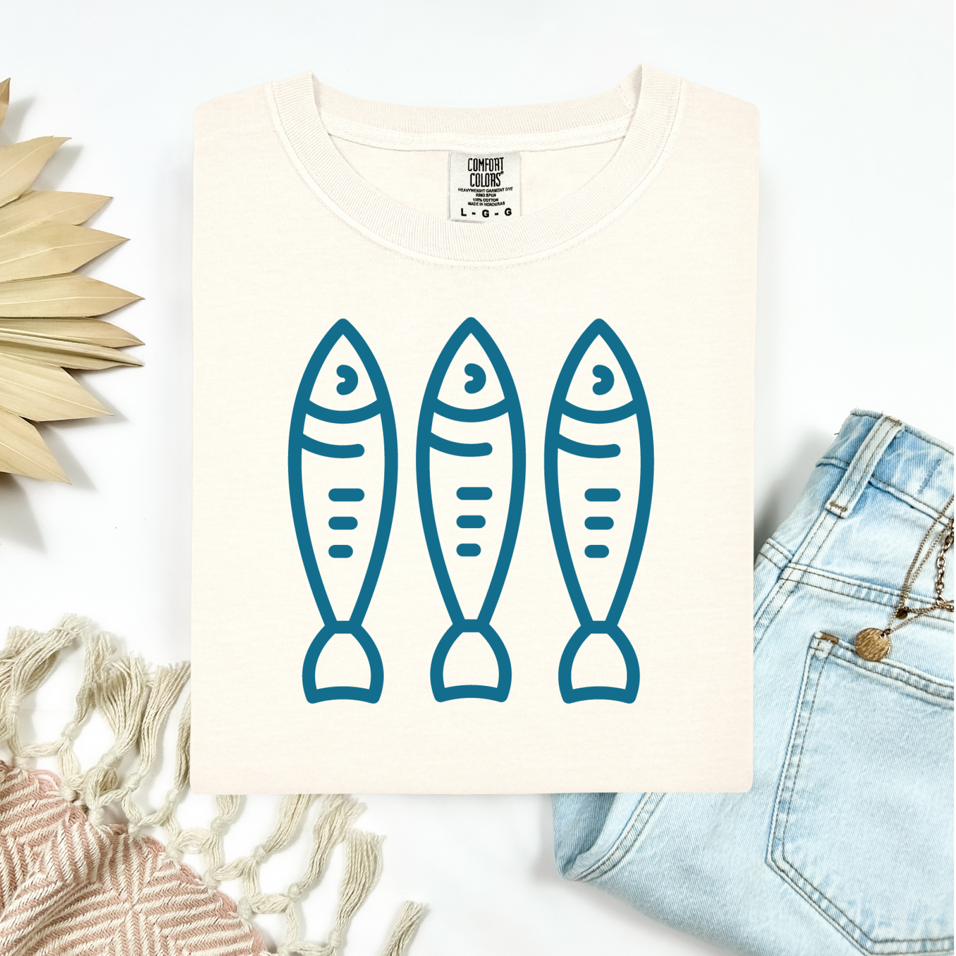 Comfort Colors 1717 Fisherman's Aesthetic t shirt with three sardines in a  one color graphic. Picture on a flay lay mockup with pair of blue jeans and blanket and a wicker sun shaped accessory.